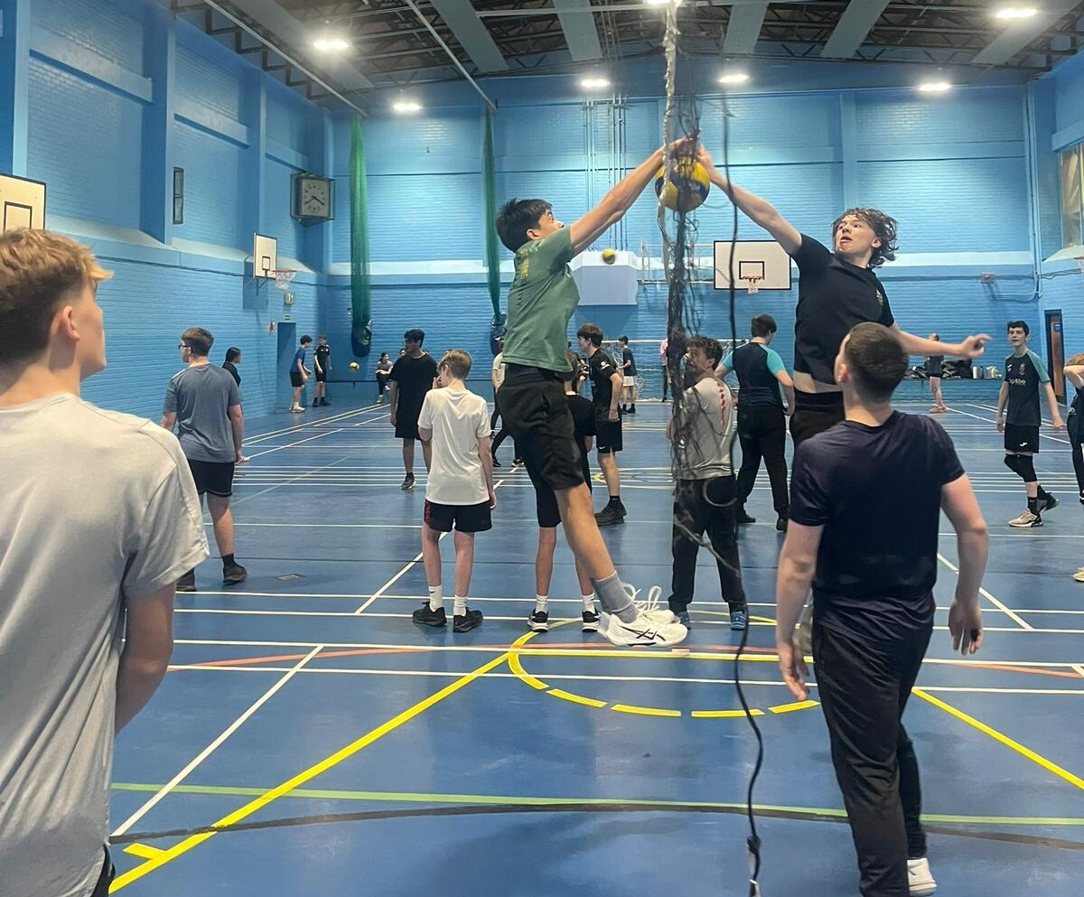 A group of high school pupils in a gym hall playing volleyball. In the foreground are two young males reaching over the net for the ball. Both the floor and the walls of the gym hall are blue.
