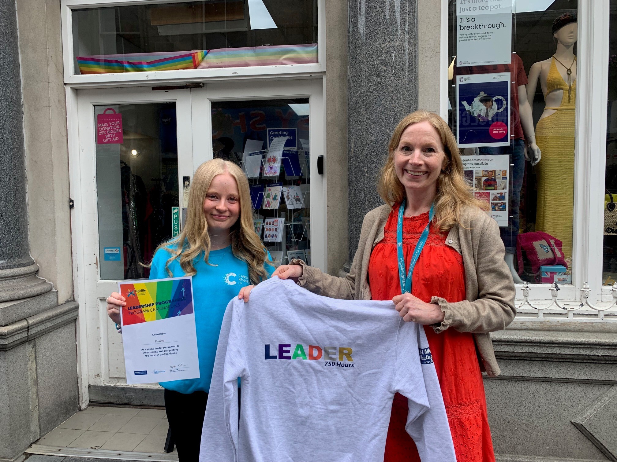 Ella White is on the left and is wearing a blue Cancer Research UK jumper and on her right hand she is holding her Leadership Programme and her left hand is holding the platinum hoodie. Ella is being presented with the grey coloured hoodie by Andrea Andrea Mackenzie, Area Sports Development Manager with High Life Highland. She is wearing a red dress and brown jacket. They are standing outside the Cancer Research Charity Shop in Inverness City Centre.