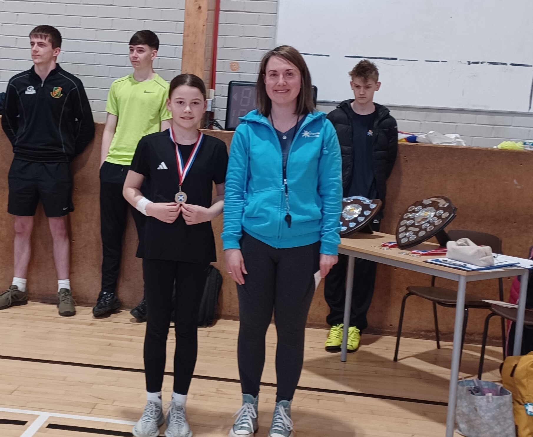 A female primary school pupil wearing dark coloured sportswear is proudly showing of her medal. She is standing beside and adult female wearing black leggings and a blue jacket. They are in a school hall setting.
