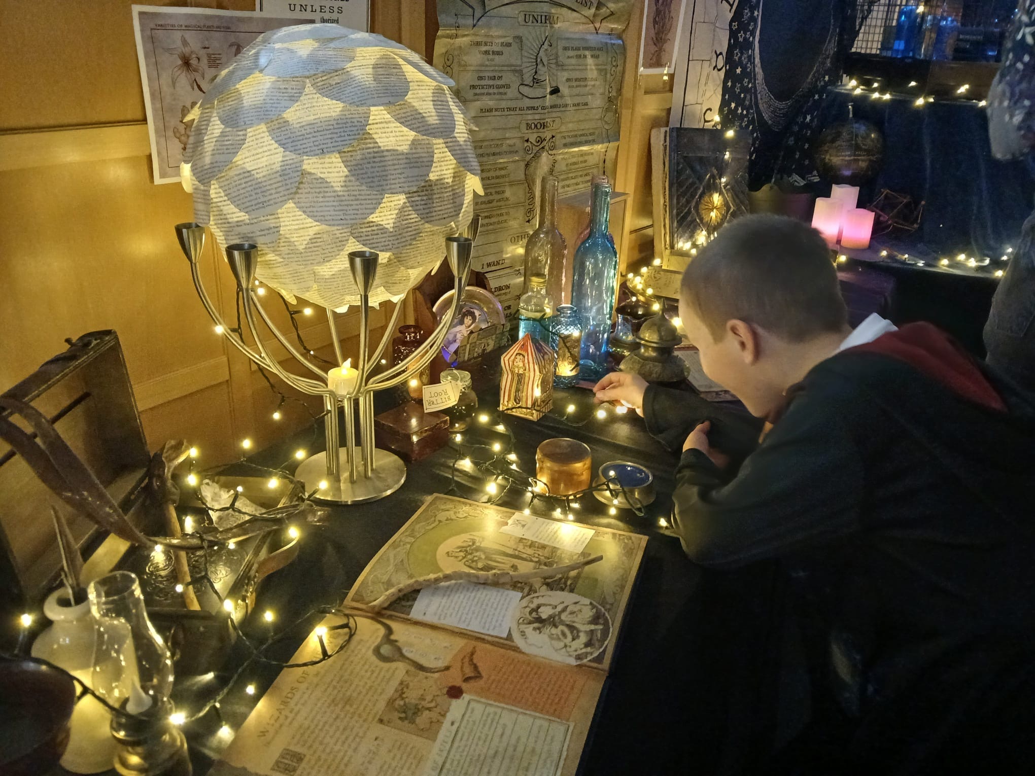 A young boy dressed as a wizard looking at a display of Harry Potter themed activities inside a library. There are lots of sparkly lights.