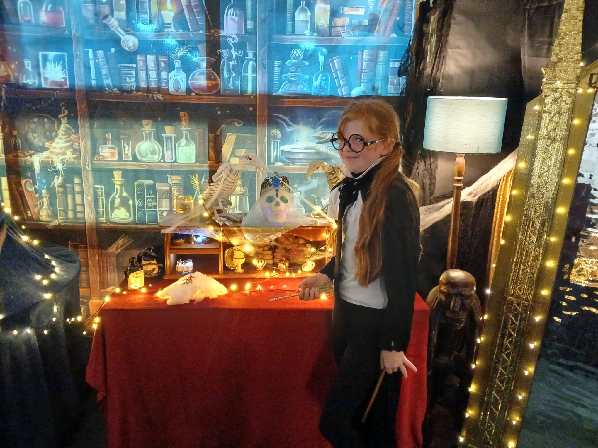 A young girl dressed as a witch with glasses on is standing by a table filled with witchcraft and wizardry items with lots of sparkly lights.