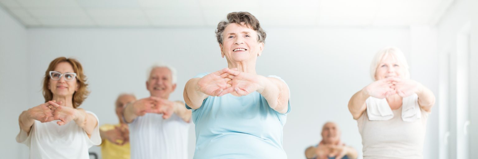 Group of older adults performing seated stretching exercises with arms extended forward in a bright fitness studio.