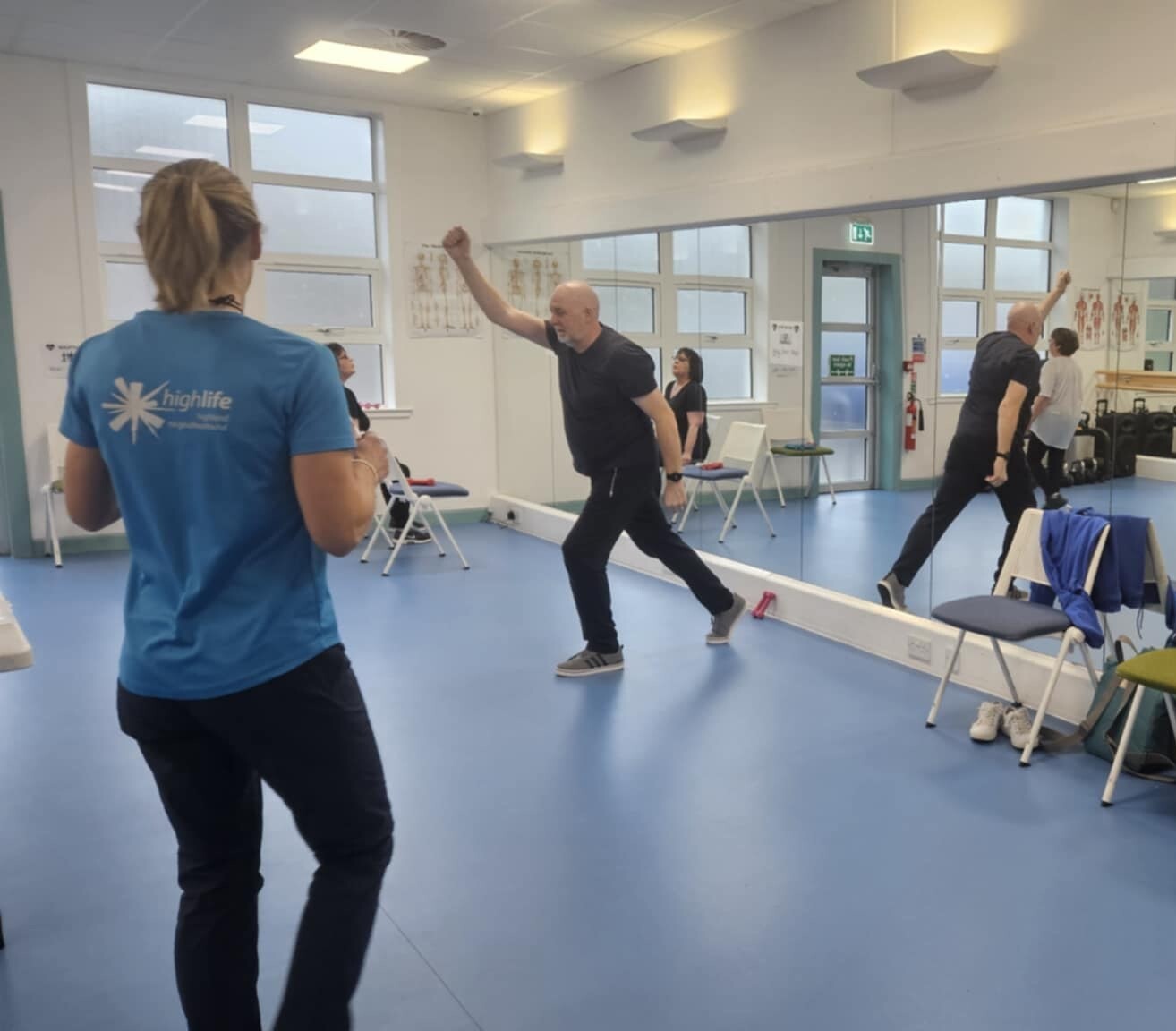 Instructor leading an exercise session in a bright fitness studio with participants performing standing movements.