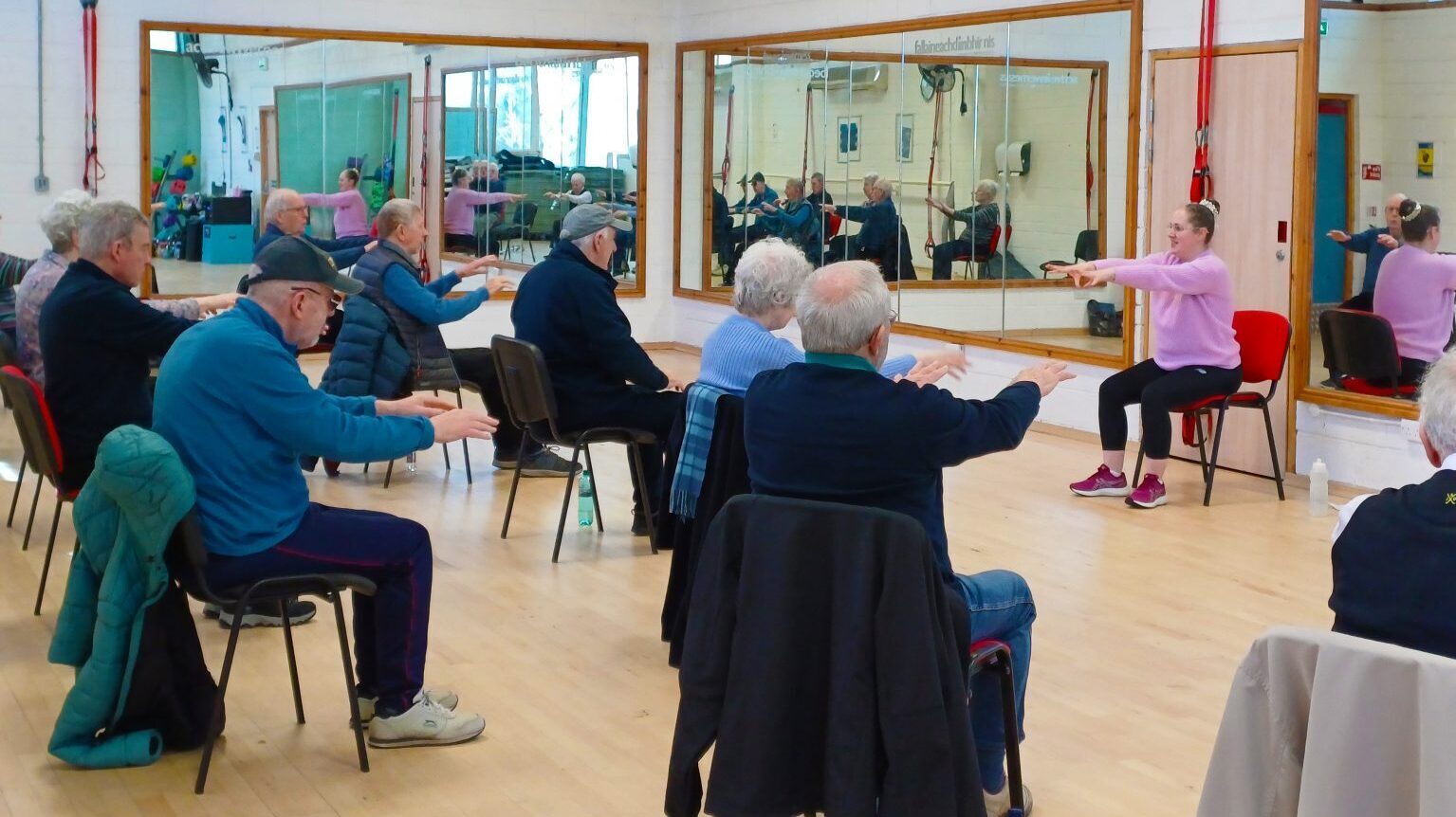 Group of older adults seated in a fitness studio following an instructor leading arm exercises in front of mirrors.