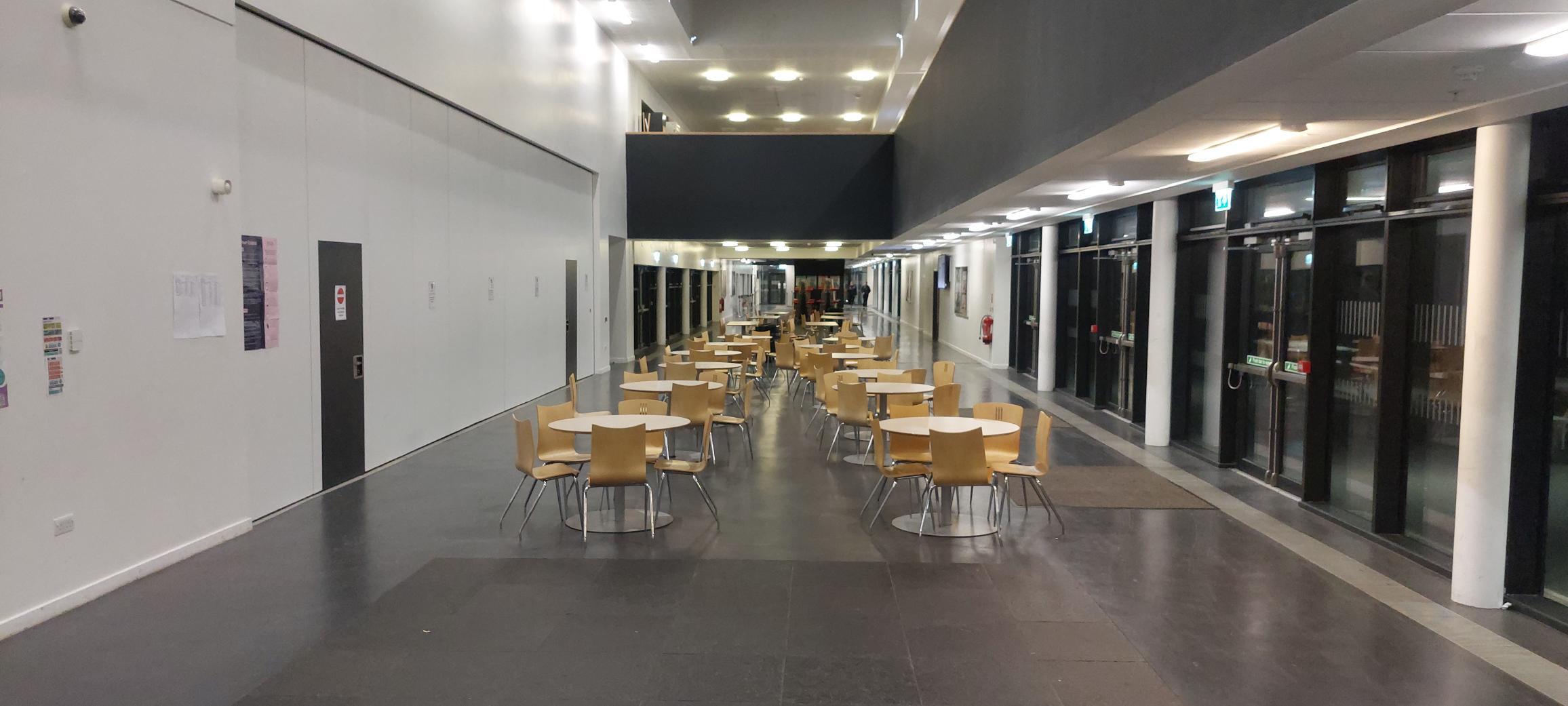 Interior of a high school corridor, nicknamed the 'street', which is wide enough for multiple round tables and chairs with floor to ceiling windows along the corridor.