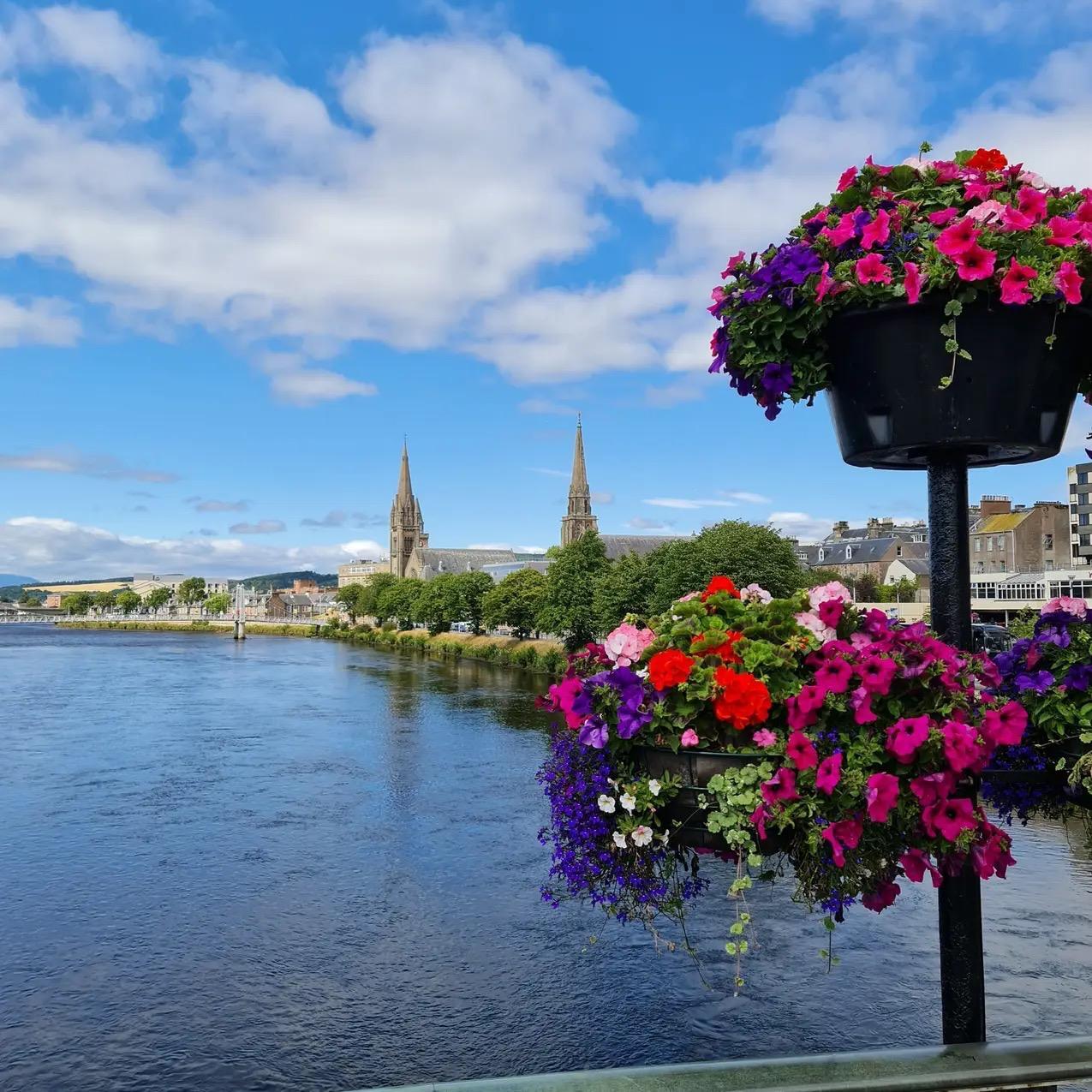 A riverside view of Inverness with Church buildings in the background and colourful hanging baskets in the foreground.