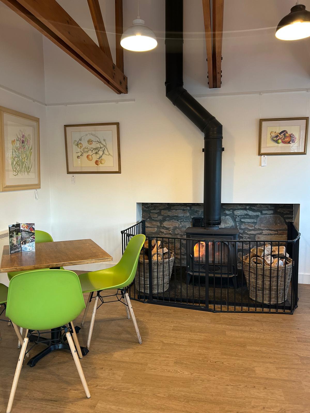 A cosy café space with a table beside a wood burning fire and paintings on the wall.