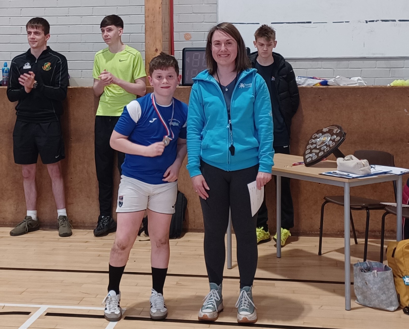 A young school pupil in white shorts and a blue top is proudly displaying his medal. He is standing next to an adult female in black leggings and a blue jacket. They are in a school hall setting.