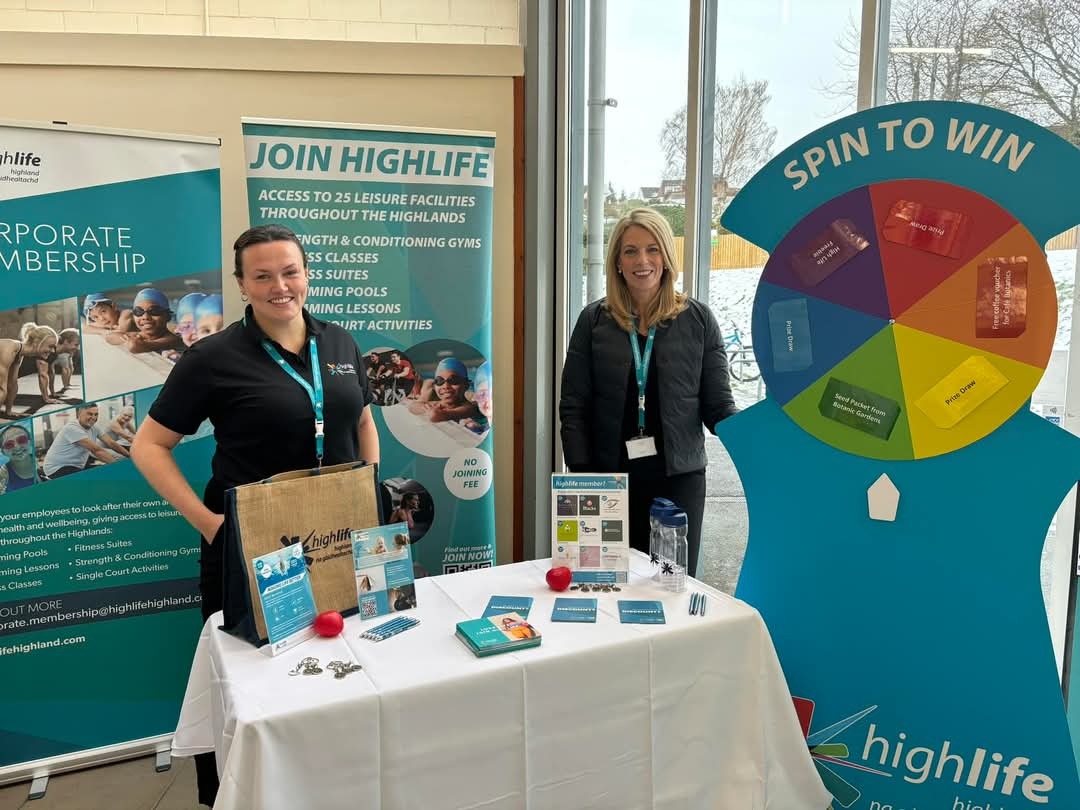Two women in black clothing wearing blue lanyards are standing behind a table with a white cloth and multiple items on it. Behind them is a Join High Life banner and a Spin to Win display.