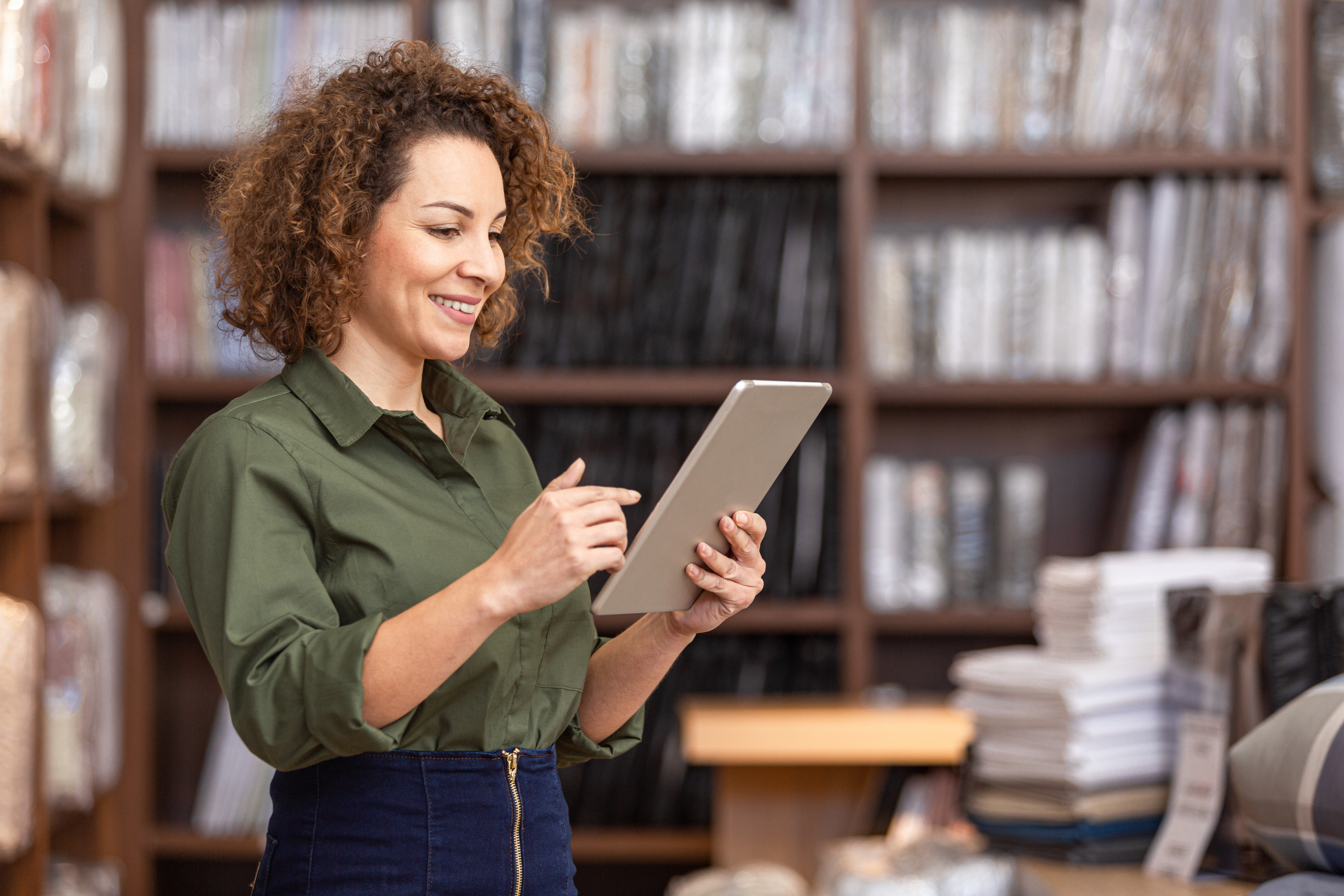A person standing in a shop or library-like space, holding and using a tablet while surrounded by shelves filled with neatly stacked items.