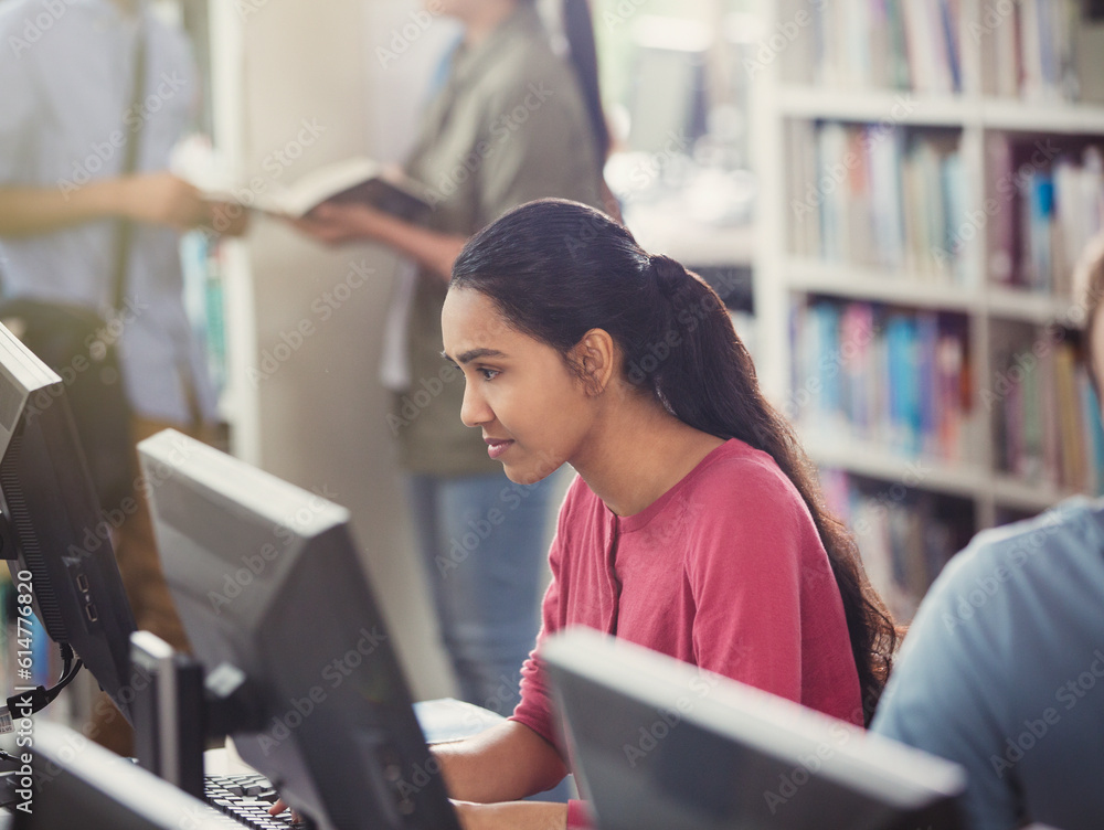 Person using a desktop computer in a library with bookshelves and other people in the background.