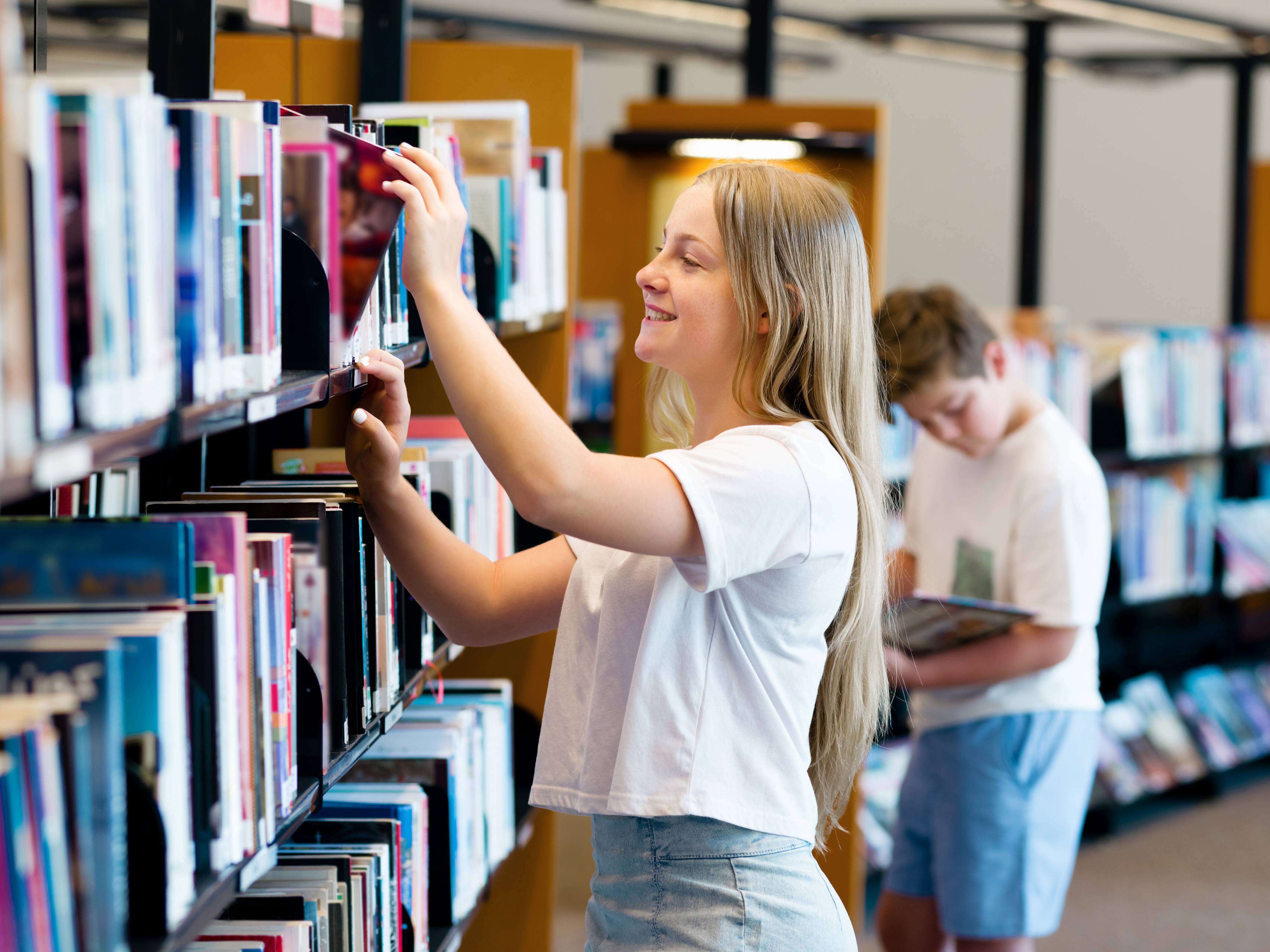A young person reaches for a book on a high shelf in a library, while another child in the background looks through a book in a different aisle. Bookshelves filled with colorful titles line the space.
