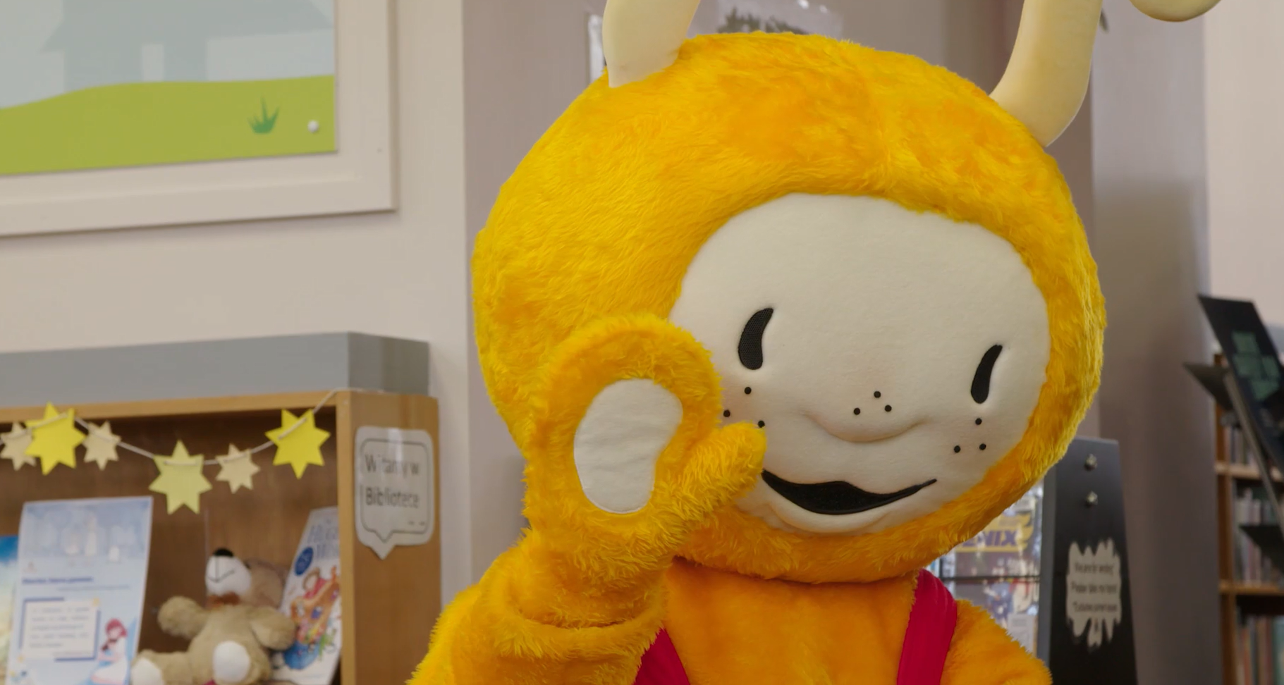 A person in a bright yellow mascot costume with antennae makes an “okay” hand gesture inside a library, with bookshelves and decorations visible in the background.