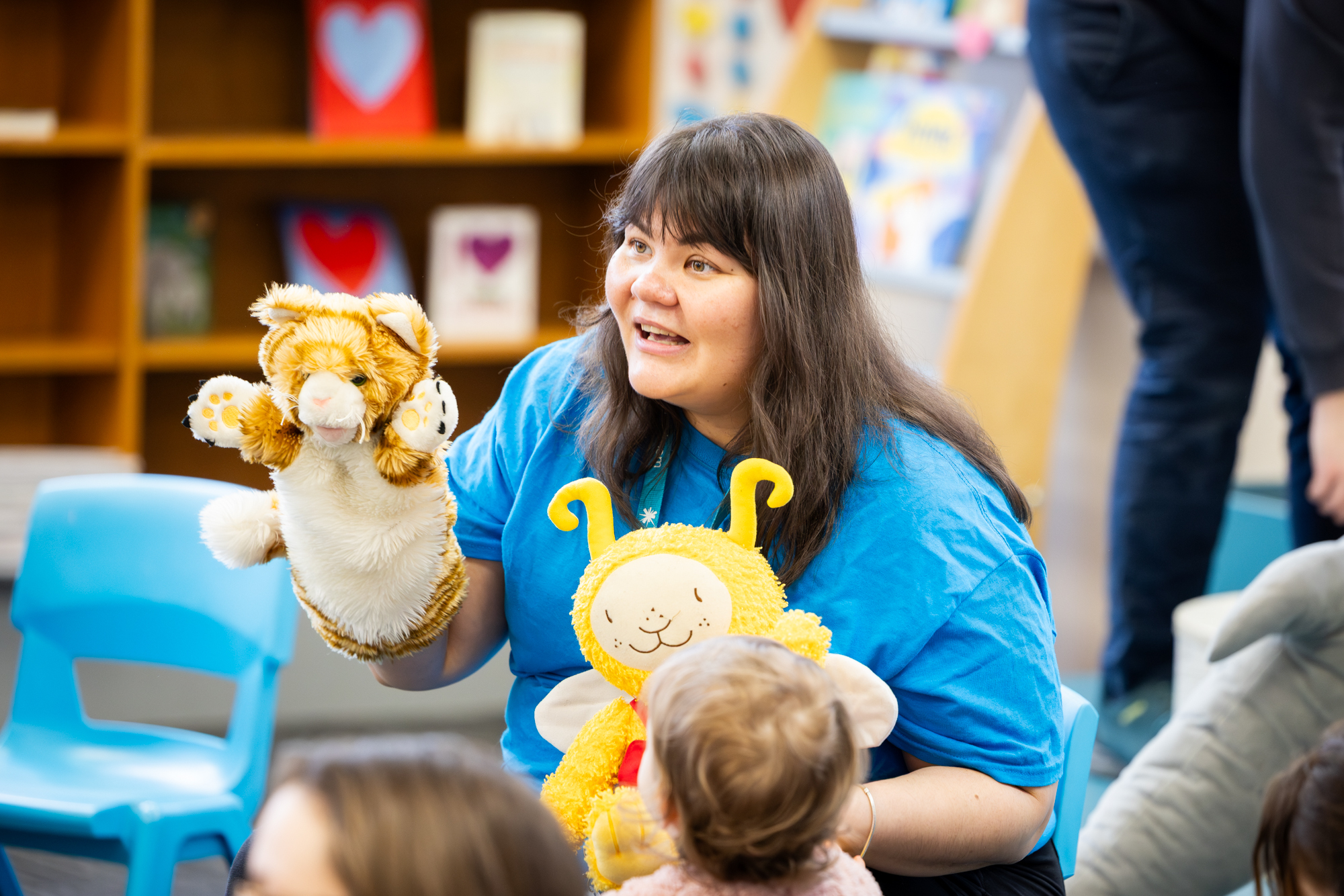 A person wearing a bright blue shirt sits in a children’s area holding two hand puppets—one resembling a tiger and the other a yellow character with antennae. A young child faces them, watching the puppets. Colourful books and small blue chairs are visible in the background.