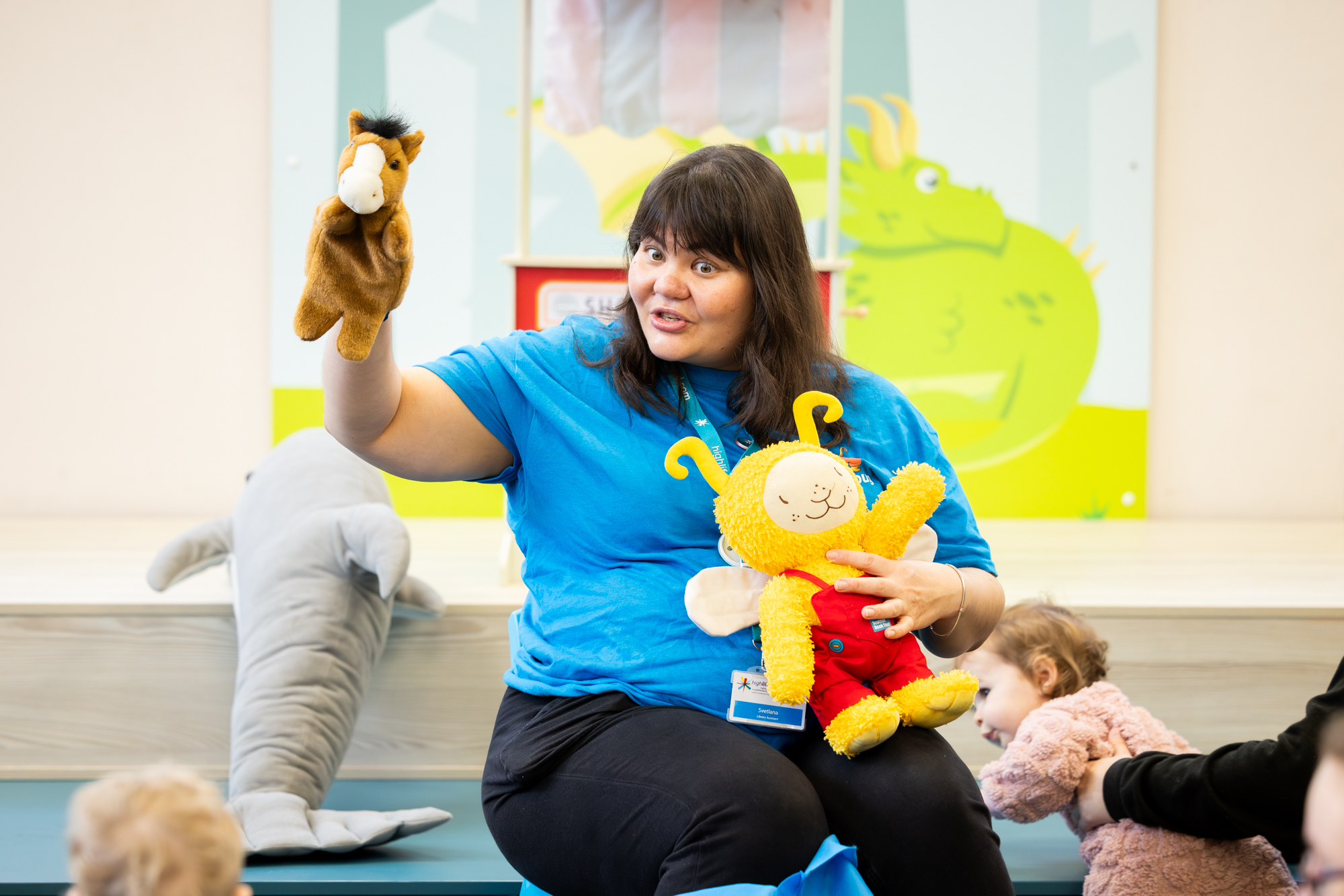 A person in a bright blue shirt sits at the front of a children’s session, holding up a horse hand puppet in one hand and a yellow plush character with antennae in the other. Several young children sit nearby watching. A large stuffed dolphin rests beside the person, and colourful illustrated wall art is visible in the background.