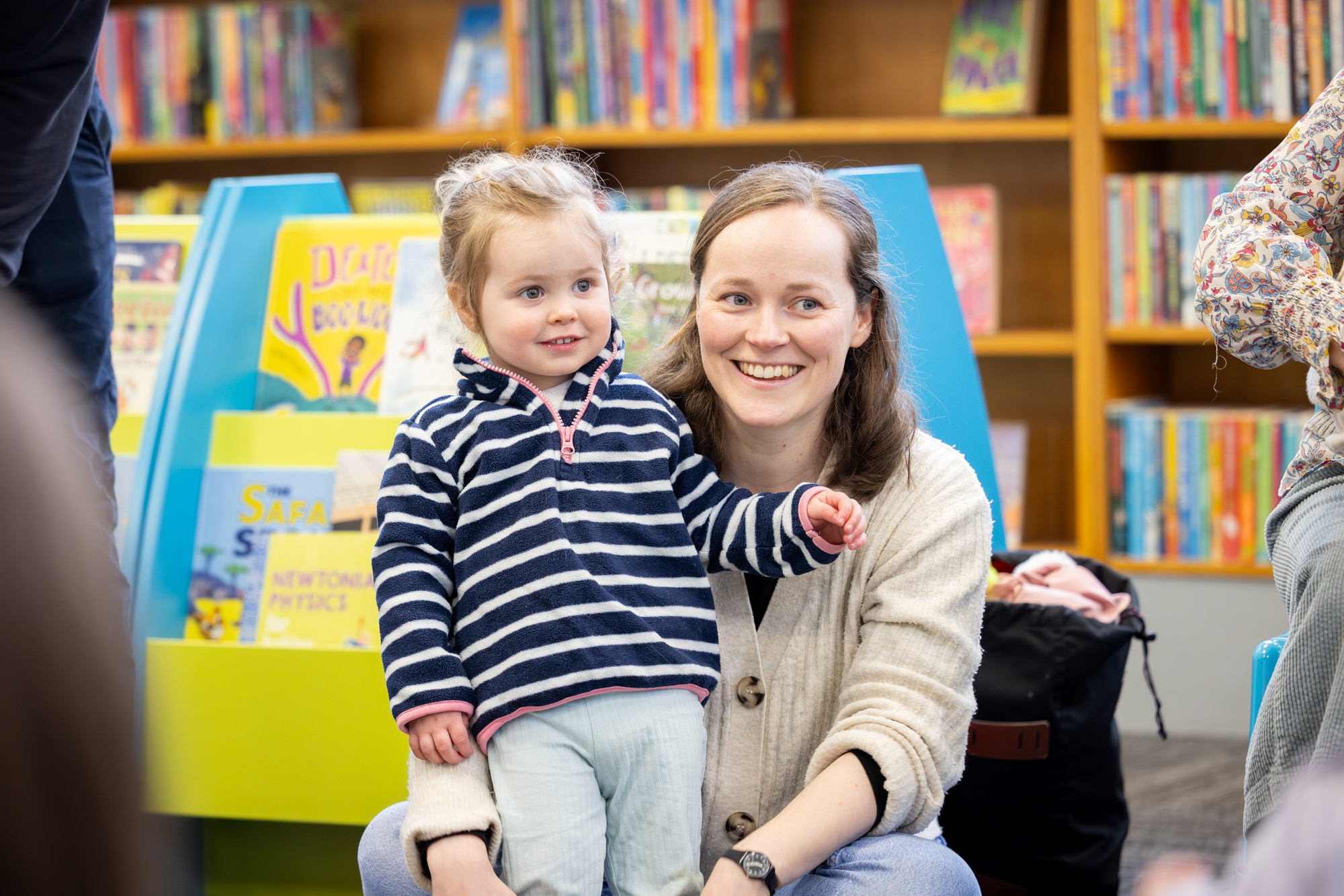 An adult sits on the floor in a library, with a child standing in front of them. Colourful bookshelves and picture books are visible in the background, and other people are seated nearby.