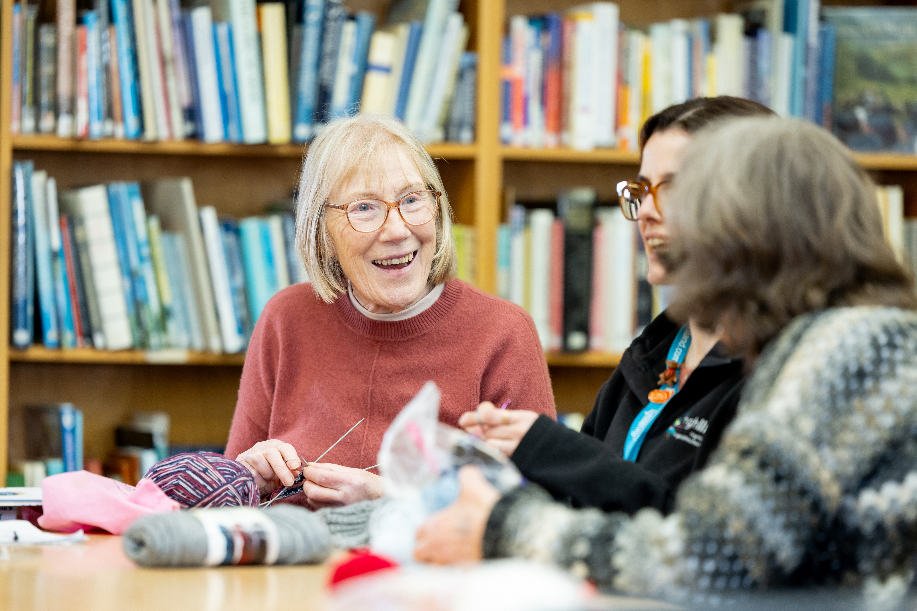 Three people sitting at a table in a library, engaged in a knitting or craft activity, with bookshelves filled with books in the background