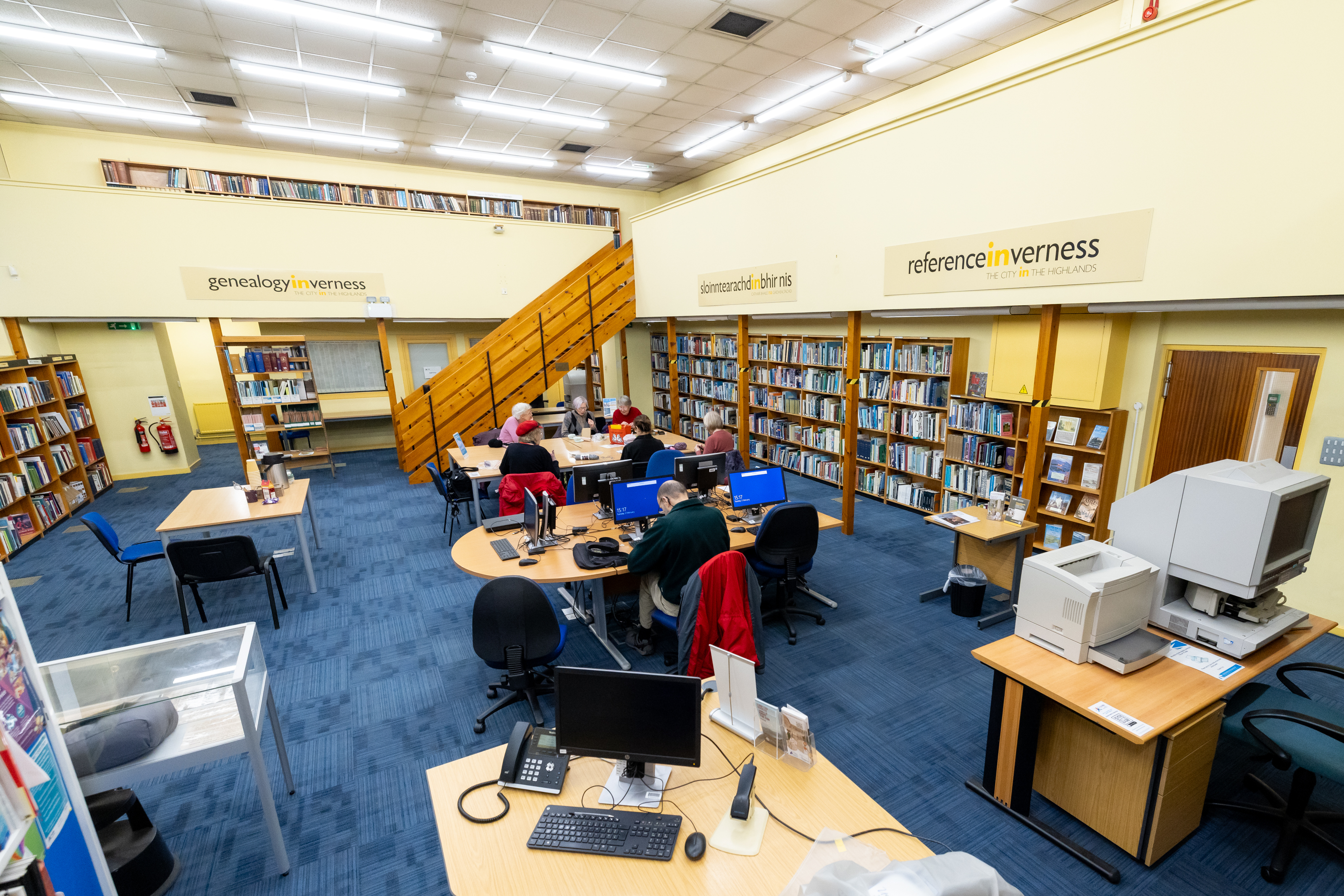 Interior of a library reference room with blue carpet, rows of bookshelves, computer workstations, and several people seated at desks using computers. A wooden staircase leads to an upper level lined with bookshelves, and signs on the walls indicate genealogy and reference sections