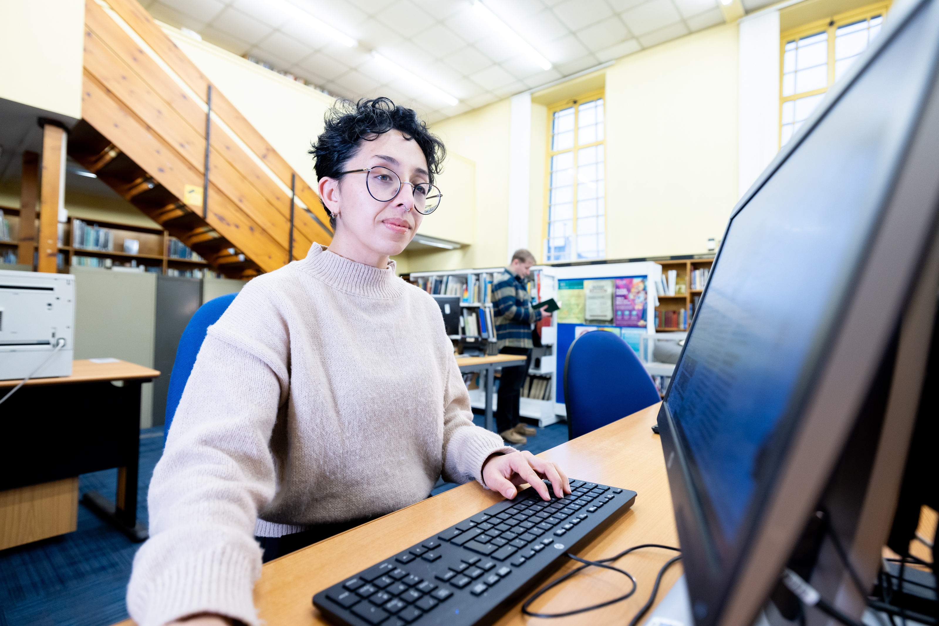 A person seated at a computer workstation in a bright library, typing on a keyboard, with bookshelves, large windows and another person browsing books in the background.