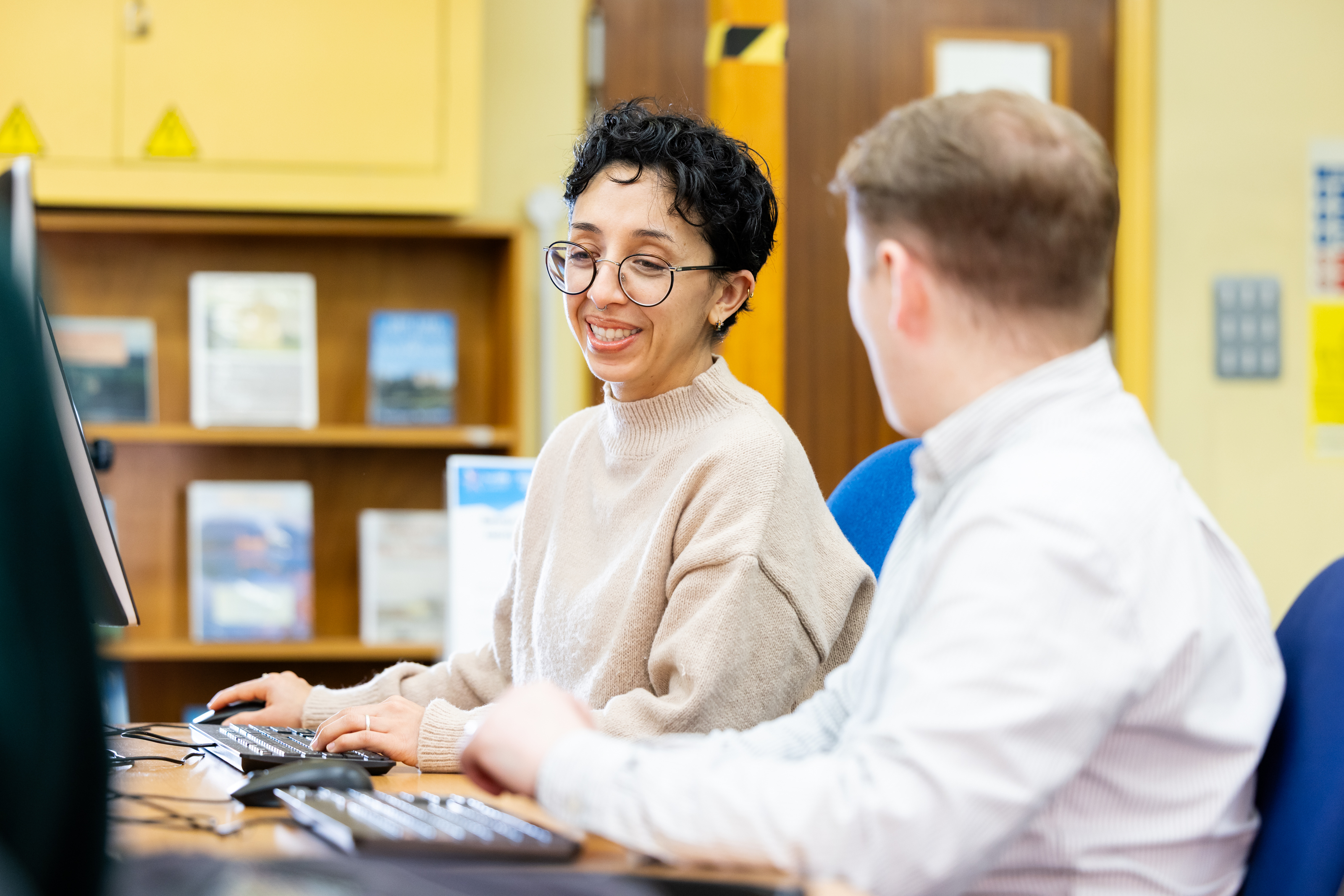 Two people seated at a computer workstation in a library, working together at a desk with bookshelves and office equipment in the background
