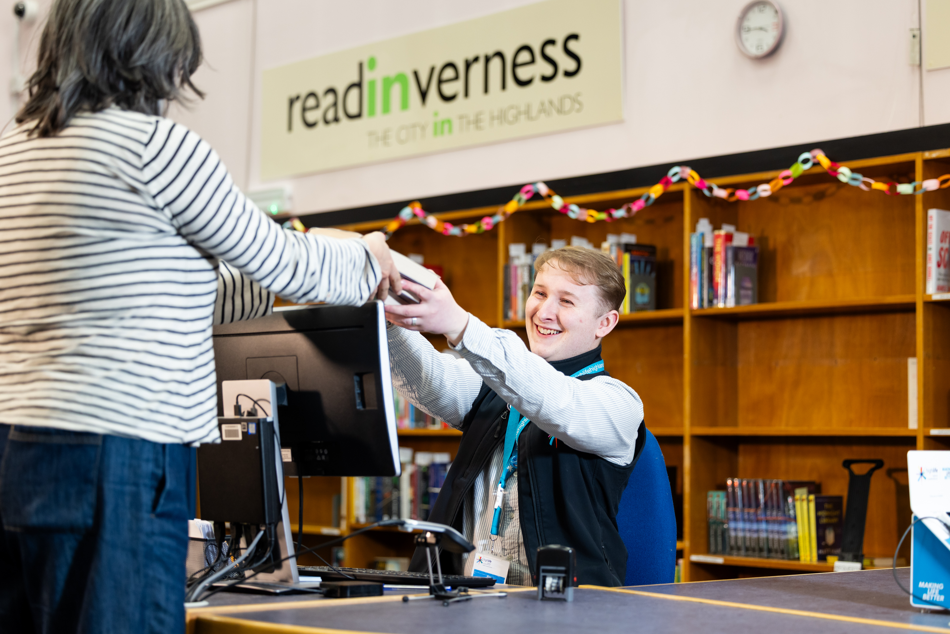 A library staff member at a service desk handing books to a visitor, with bookshelves, a computer monitor, and a ‘read Inverness’ sign in the background