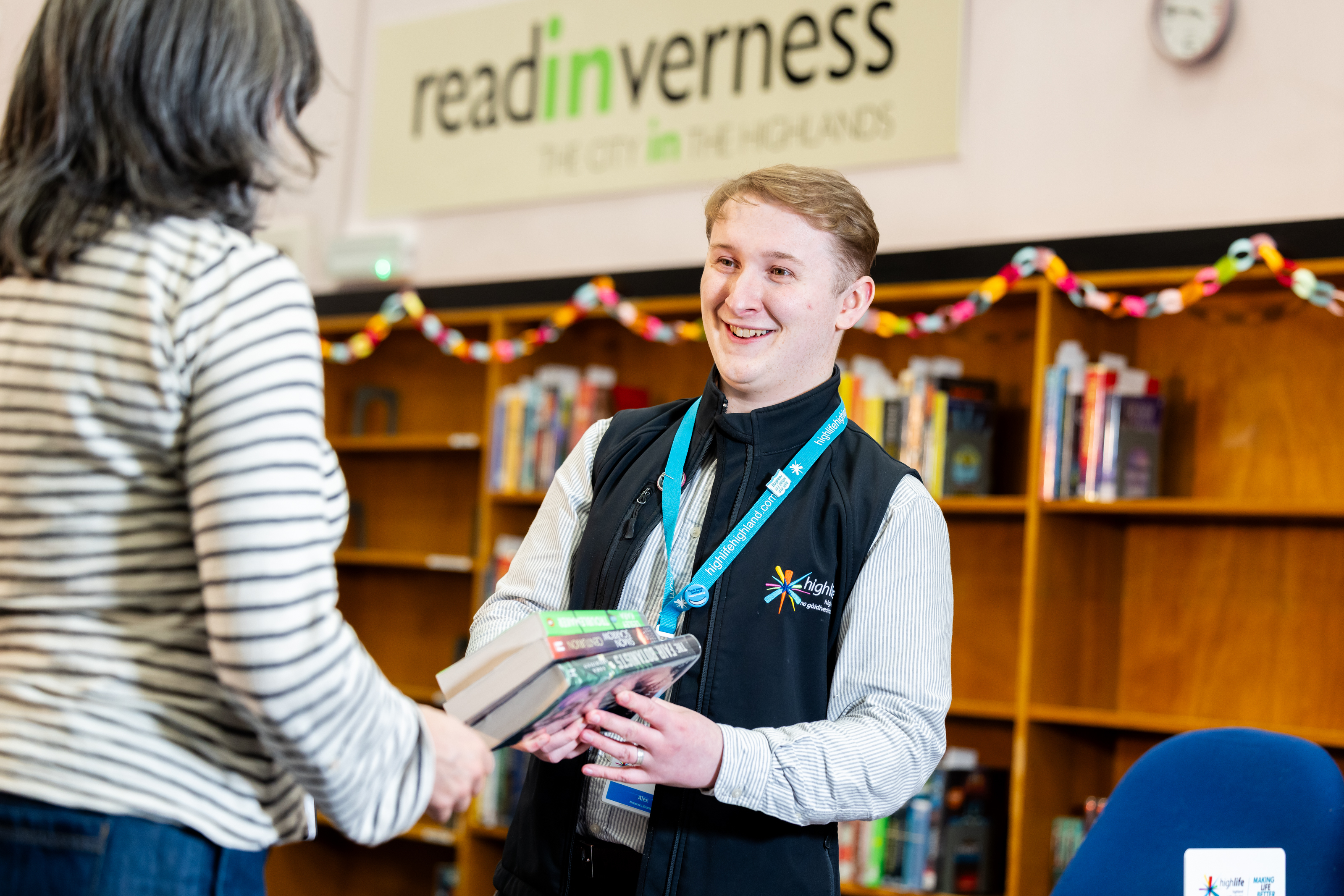 A staff member handing books to a visitor inside a library, with shelves of books and a ‘read Inverness’ sign in the background