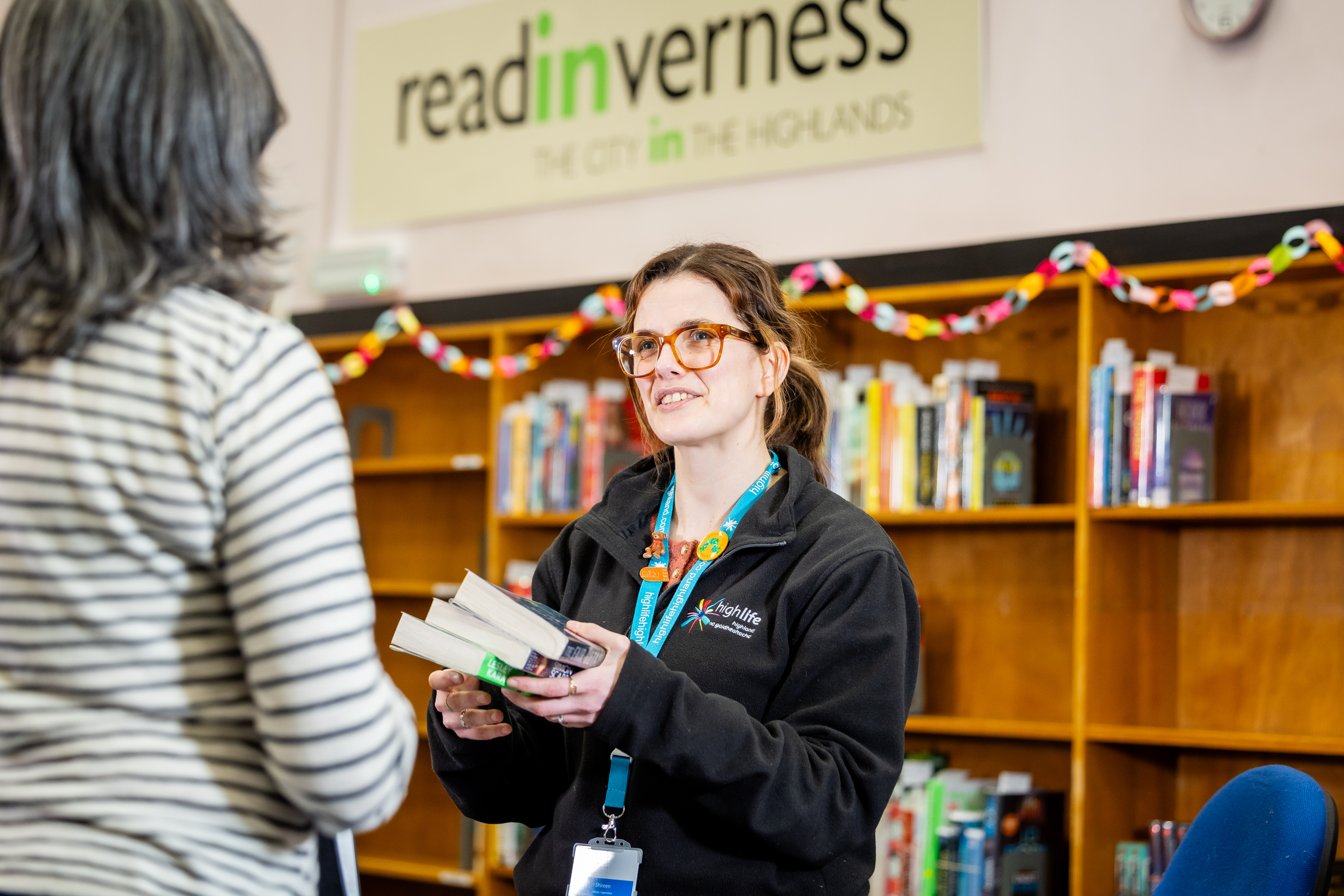 A library staff member holding several books while speaking with a visitor, with bookshelves and a ‘read Inverness’ sign in the background