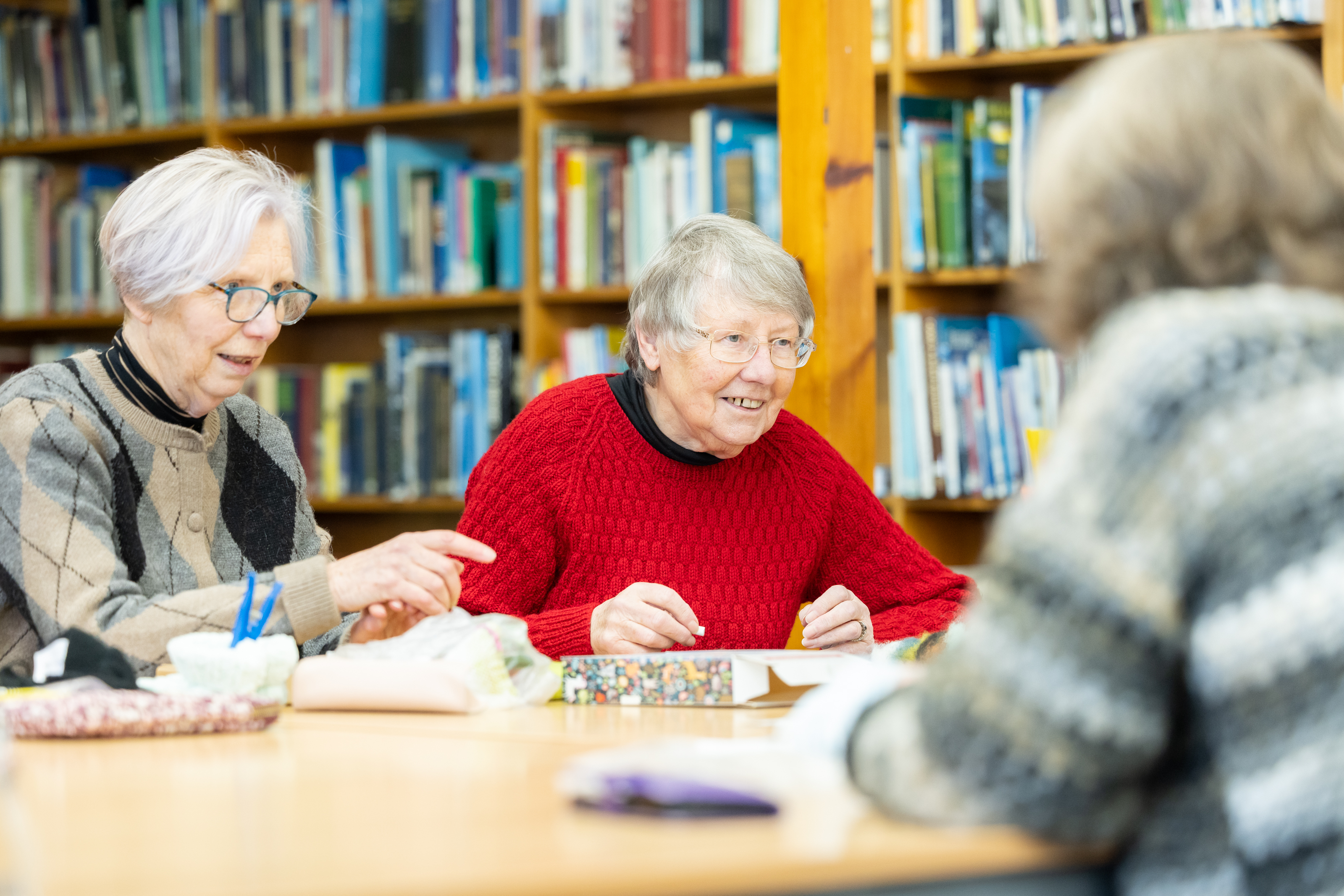 Three adults sit together at a table in a library, engaged in a craft or activity involving papers and small items. Bookshelves filled with books line the background, and the group appears to be working collaboratively in a relaxed setting.