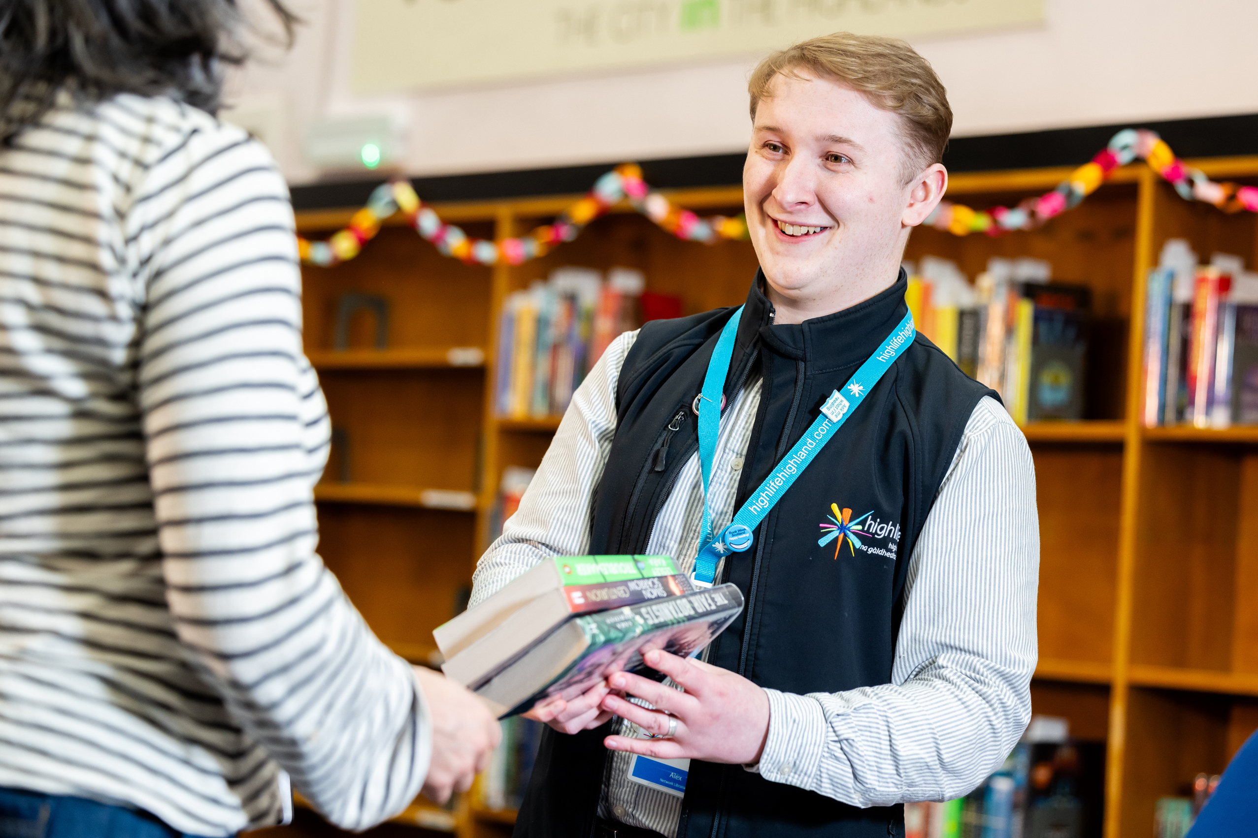 A library staff member wearing a lanyard and uniform vest holds a stack of books while interacting with a person in a striped shirt. They are standing in front of bookshelves decorated with colourful paper chains.