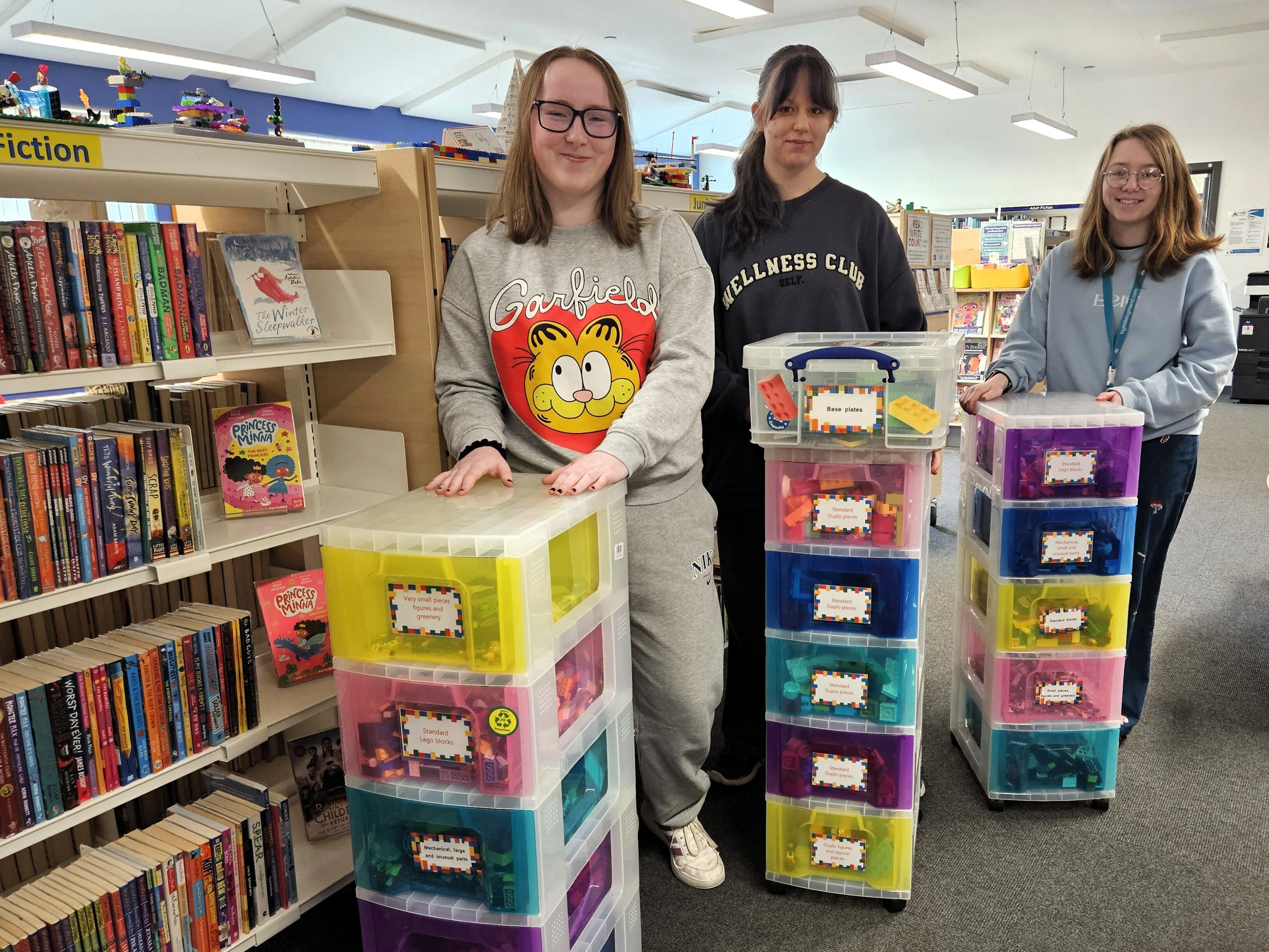 Three people stand in a library beside stacks of colourful LEGO drawer units, with shelves of books behind them and storage boxes.