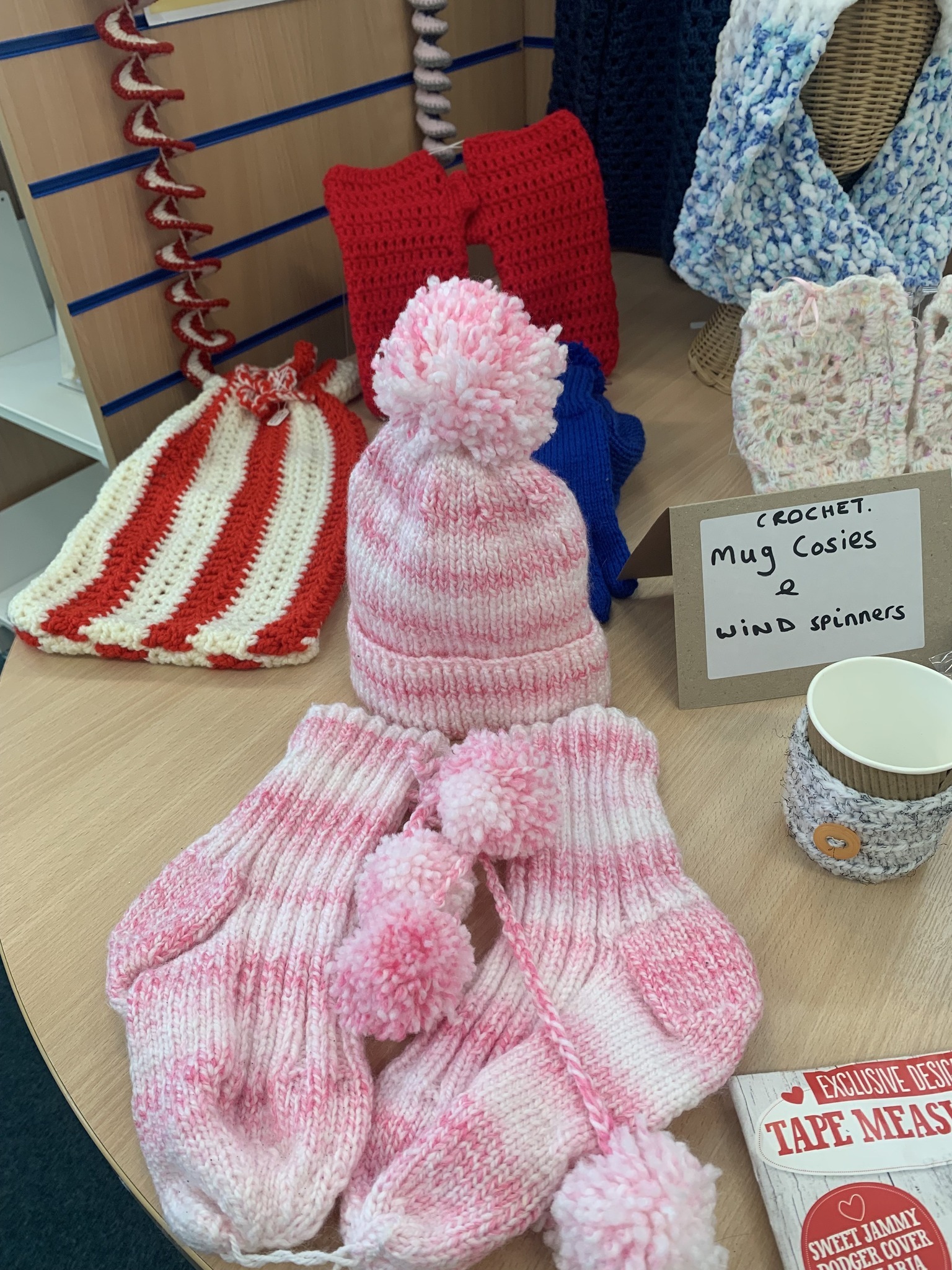 A display table with various crocheted items, including a pink-and-white striped hat with matching long mittens, a red-and-white striped piece, several red and blue rectangular pieces, and a cream and pink square motif. A small handwritten sign reads “Crochet: Mug Cosies & Wind Spinners.” A white mug with a crocheted cosy sits beside the sign, and a book titled “Tape Measure” is partly visible in the corner.