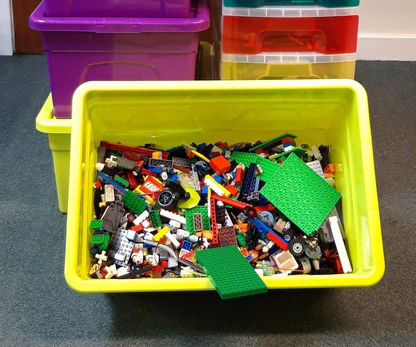 Bright yellow bin filled with assorted LEGO pieces in front of two stacked purple tubs and a tall rainbow-colored drawer unit, set against a wooden door.
