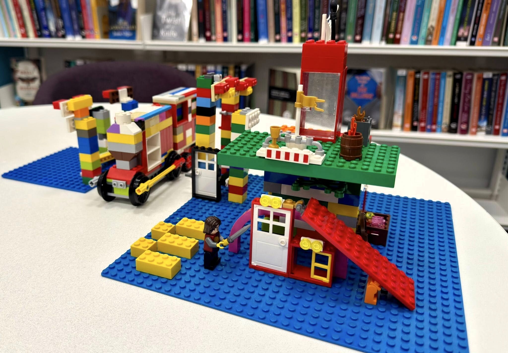 Colorful LEGO creations on blue baseplates, including a small house with doors, windows, and a figure, set on a table in a library.