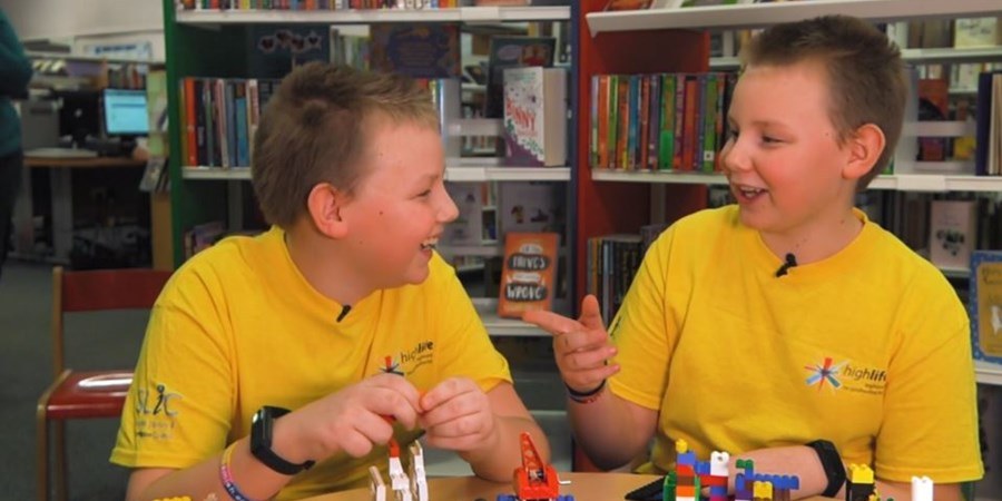 Two boys smile at each other while sitting at a table. They are wearing yellow t-shirts and playing with lego.