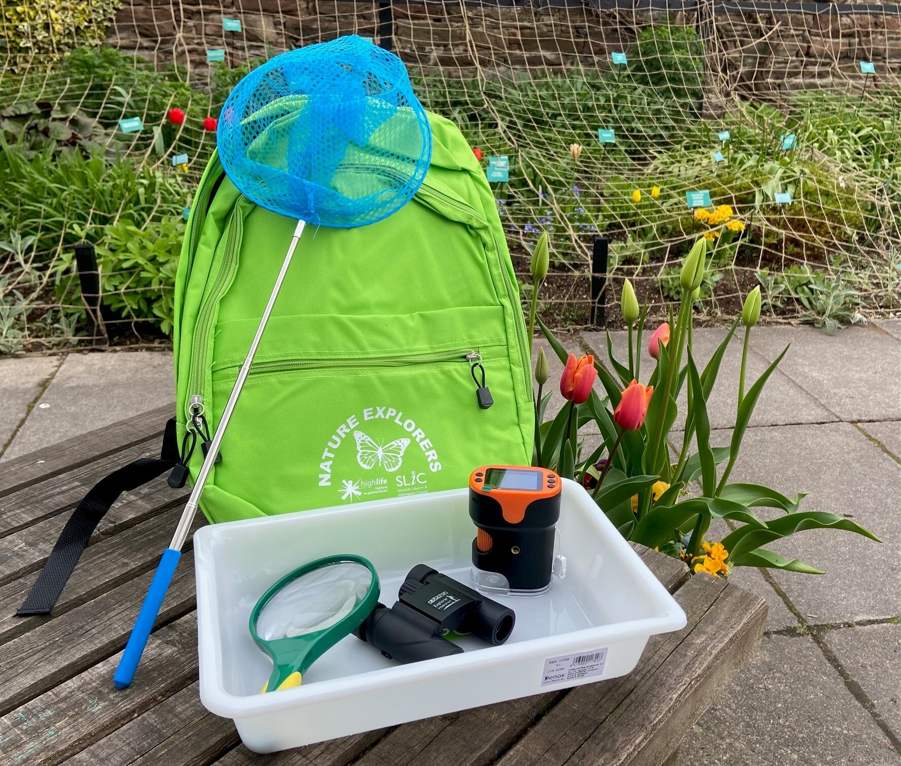 Green Nature Explorers backpack with a net, magnifying glass, binoculars, and a small microscope in a tray, placed outdoors beside blooming tulips.