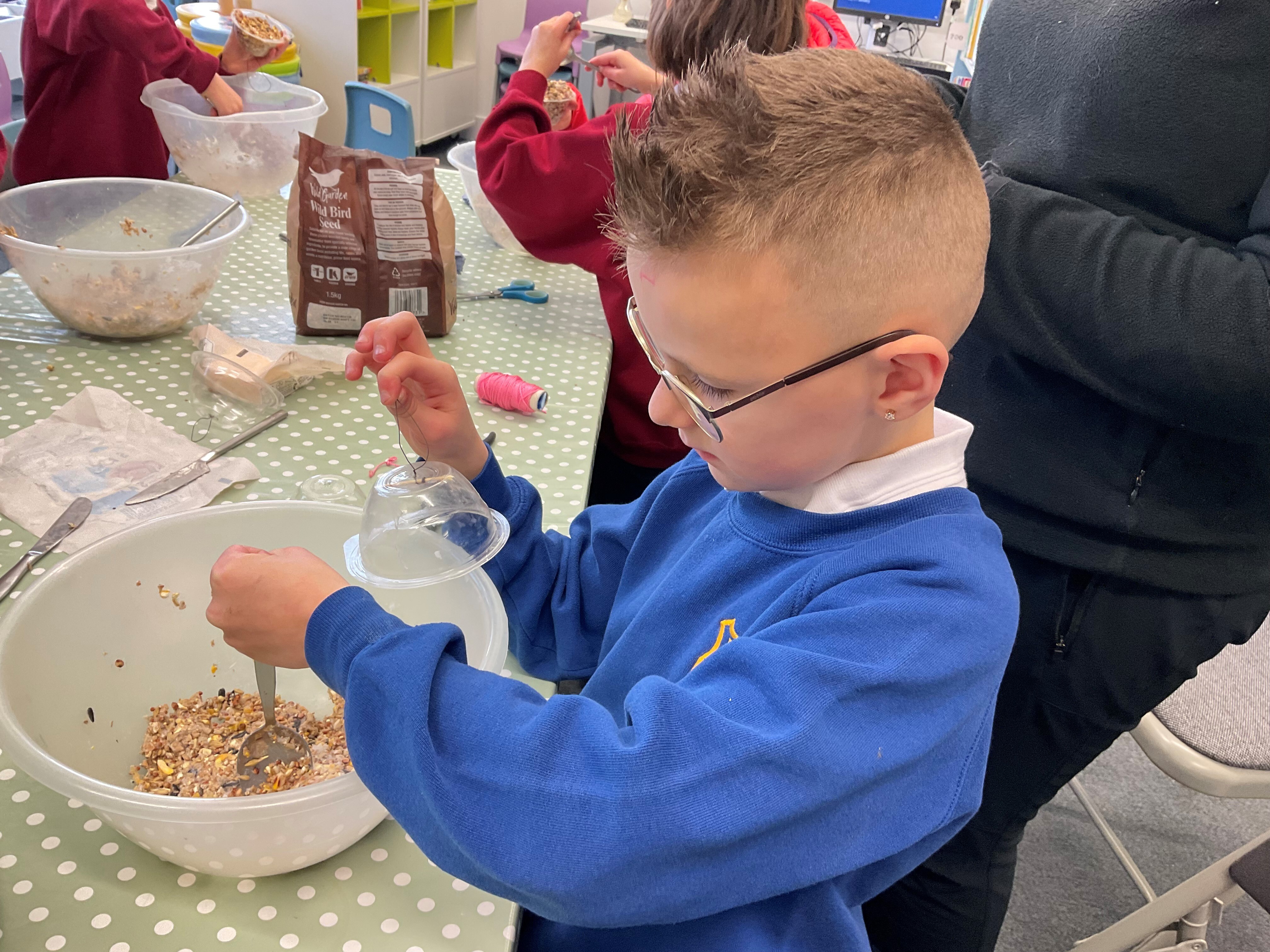 A boy in a blue jumper and wearing glasses is sitting at a table making bird food. He is mixing ingredients in a large plastic bowl.