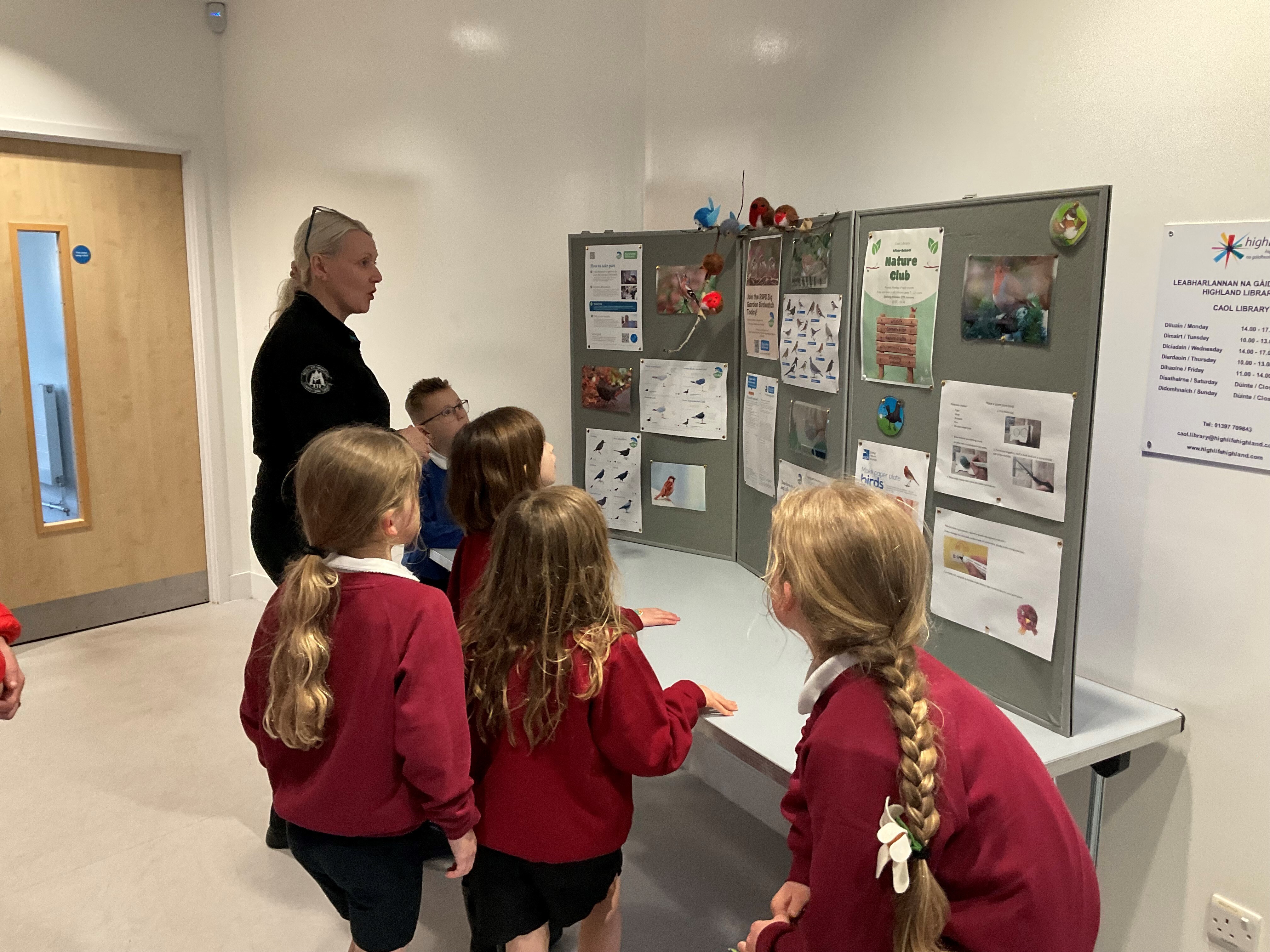 A woman in a dark jumper with light hair is standing looking at a nature display board in a library setting. She is accompanied by 5 children in red jumpers.