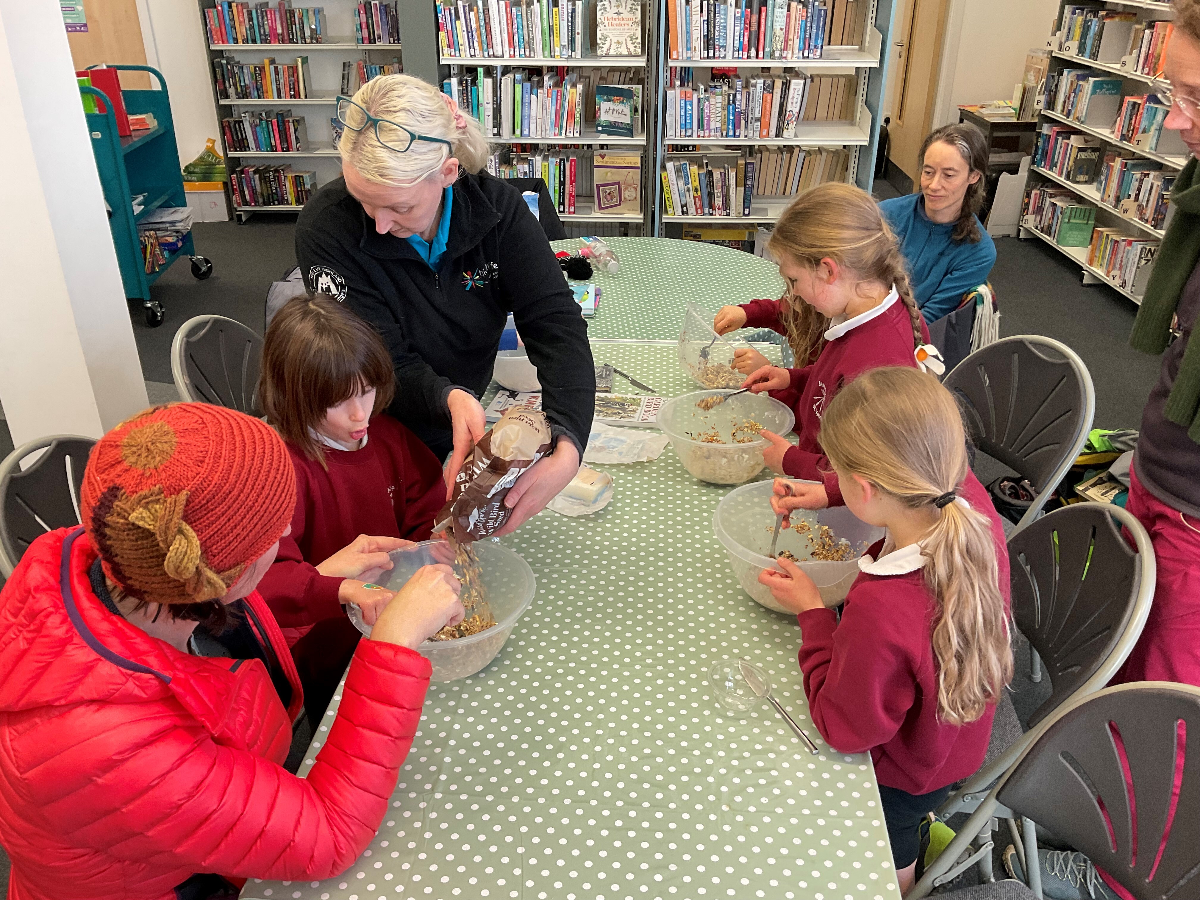 A woman in a dark jumper with blonde hair is showing a group of three children in red jumpers, along with two other adults, how to make homemade bird food. They are sat at a long table with a green tablecloth within a library setting.