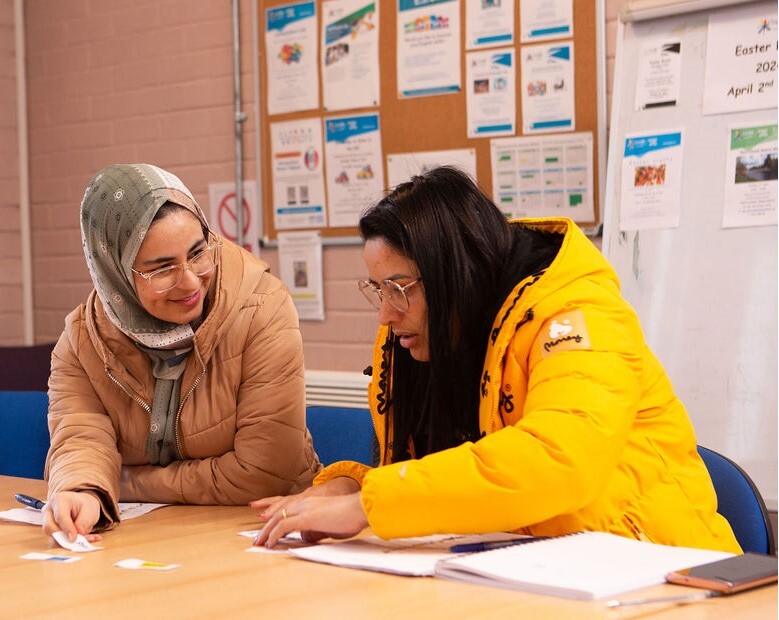 Two people seated at a table in a community room, working together on papers, with noticeboards and a flip chart in the background.