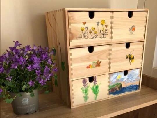 Small wooden drawer unit with hand‑painted nature designs, placed on a wooden shelf beside a pot of purple flowers.