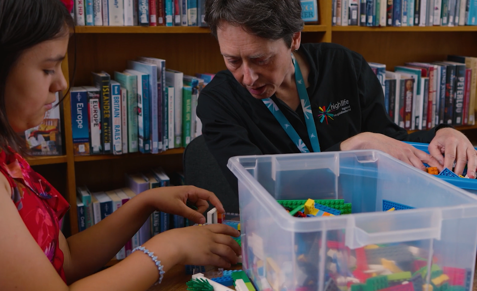 Two people sitting at a table in a library, sorting and building with colourful LEGO bricks from a large plastic container, with bookshelves in the background
