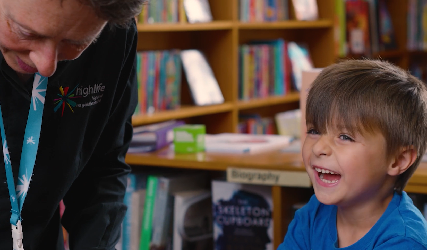 A staff member wearing a High Life Highland lanyard leans over to assist a child in a library, with bookshelves filled with colourful books in the background.
