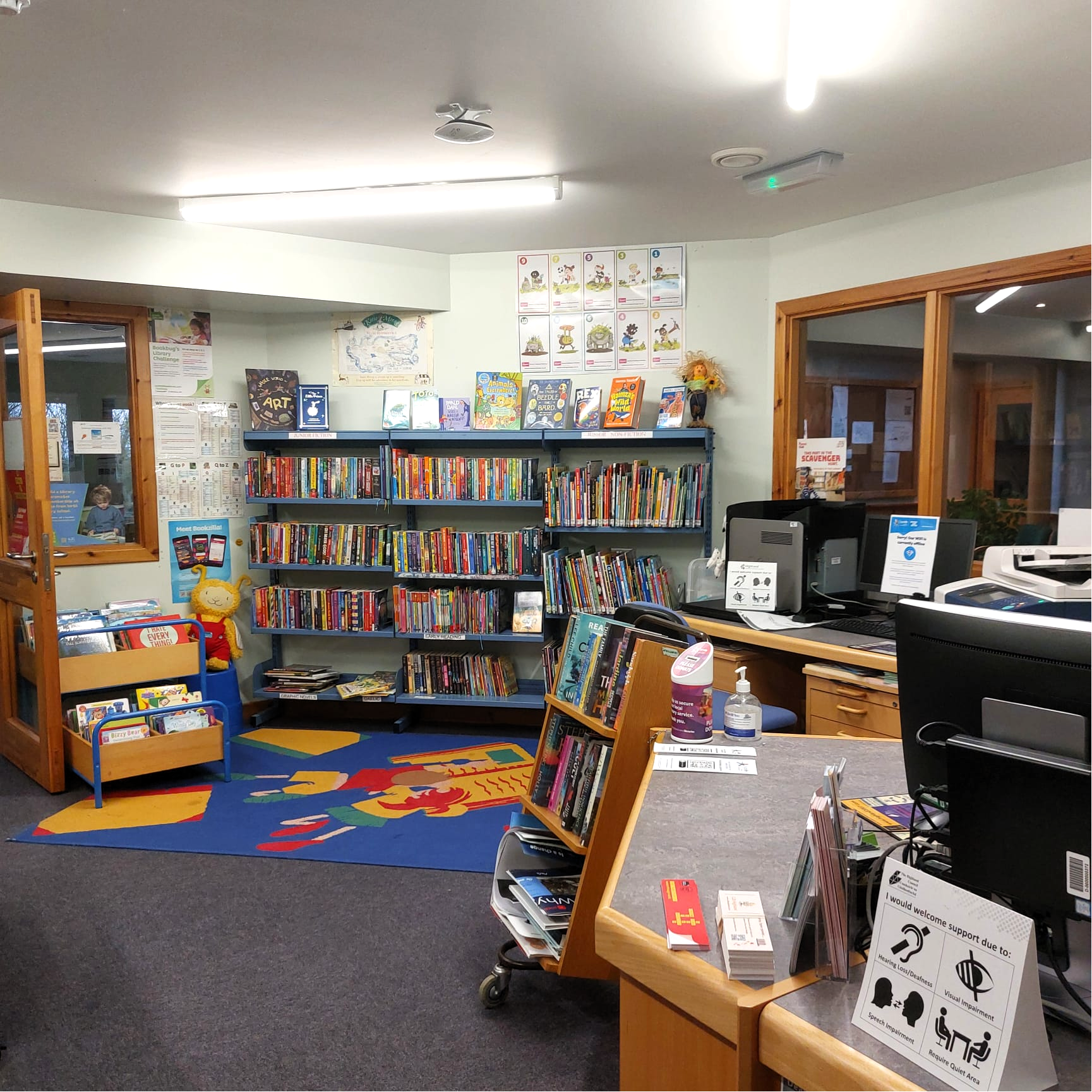 Children’s area in a small library with bookshelves, a colourful rug, and a service desk with computers.