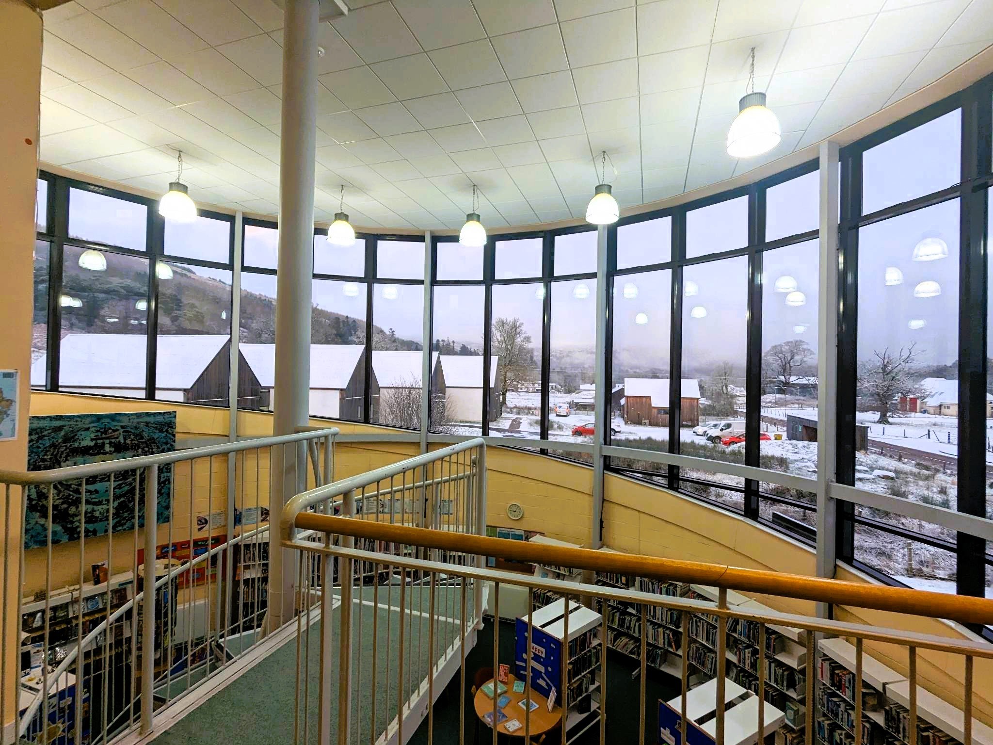 Interior view of a modern library with large curved floor-to-ceiling windows overlooking a snowy landscape. The windows reveal snow-covered rooftops, hills, and trees outside. Inside, there are hanging ceiling lights, a metal railing around an upper level walkway, and bookshelves visible on the lower floor.
