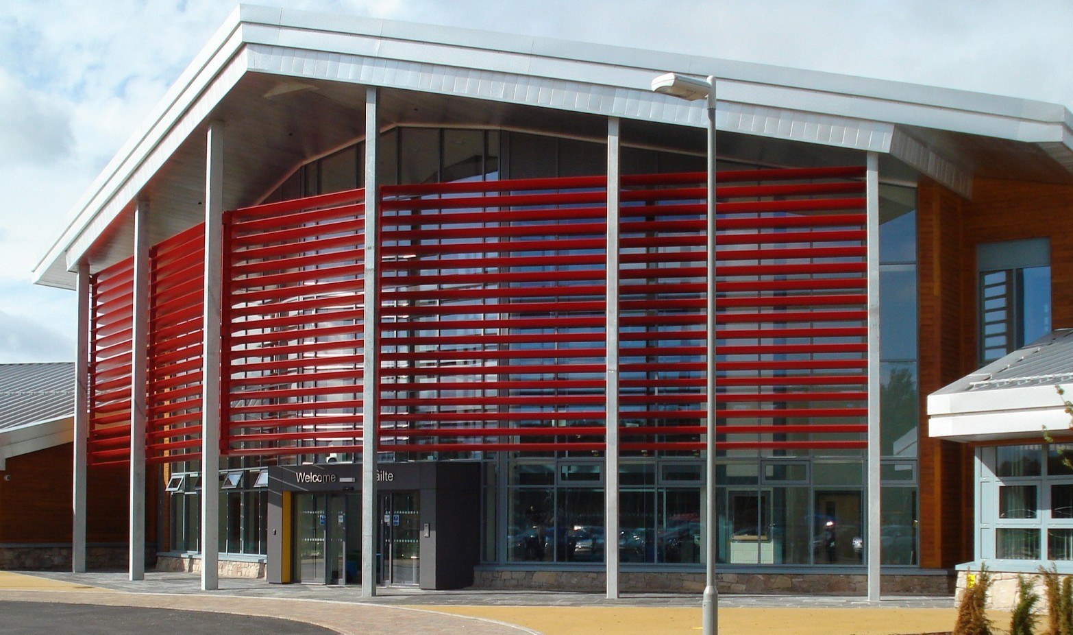 Modern building with a glass façade partially covered by horizontal red slats, tall silver columns, and a sloping metal roof.