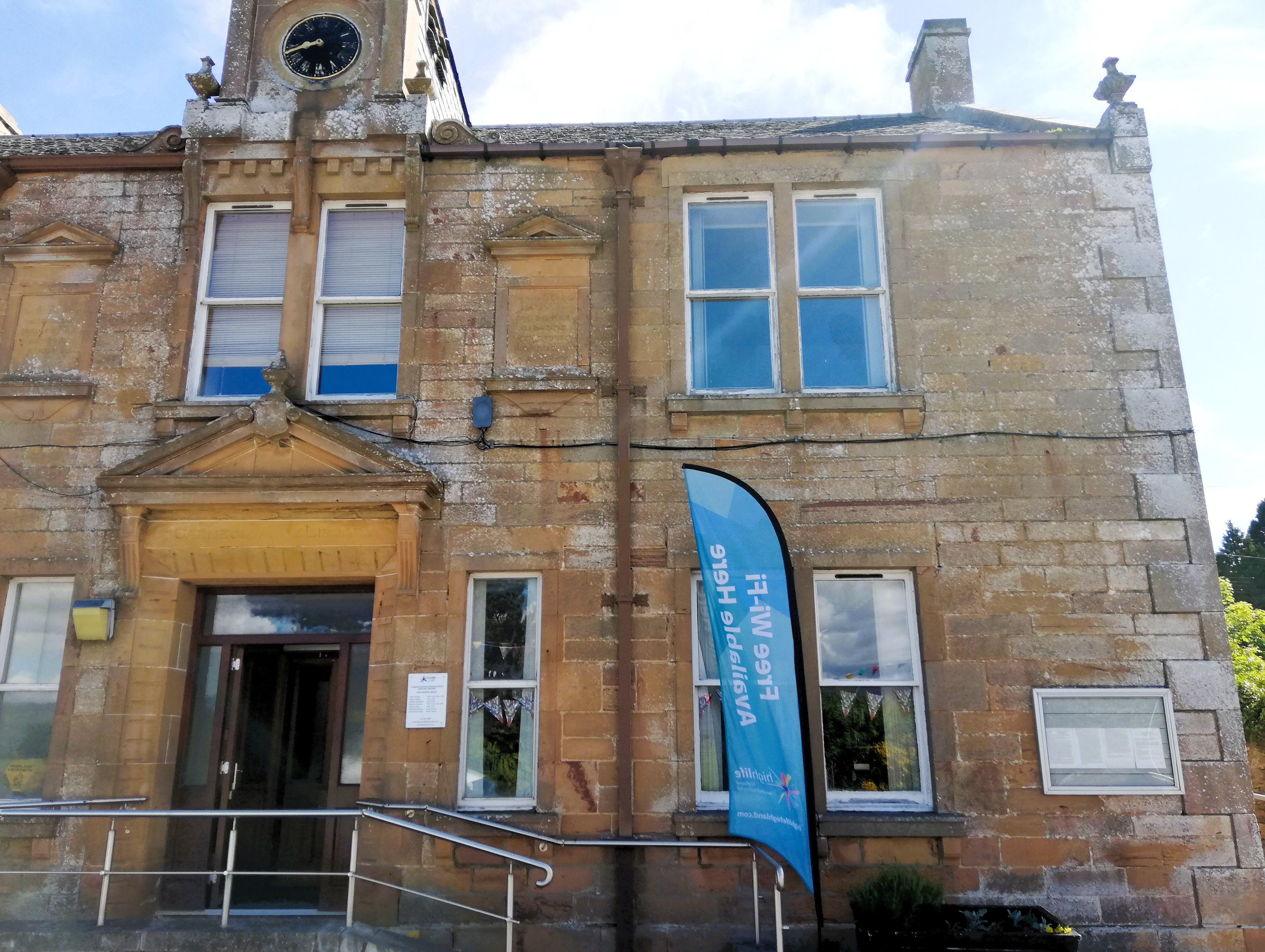Front view of Bonar Bridge Library, a sandstone building with a clock tower at the top center. The entrance has a ramp with metal railings leading to a wooden door under a triangular pediment. A blue vertical banner with white text stands near the doorway, and several windows are visible on the upper and lower floors.