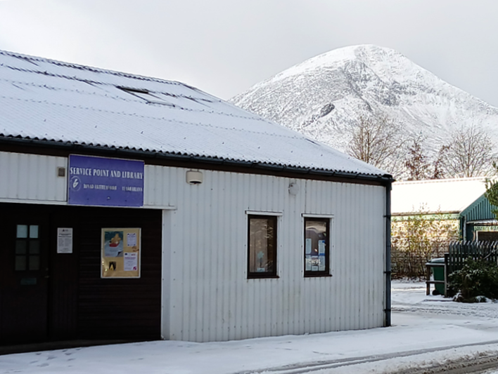 Snow-covered buildings and parked cars with a tall, snowy hill rising in the background on a winter day.