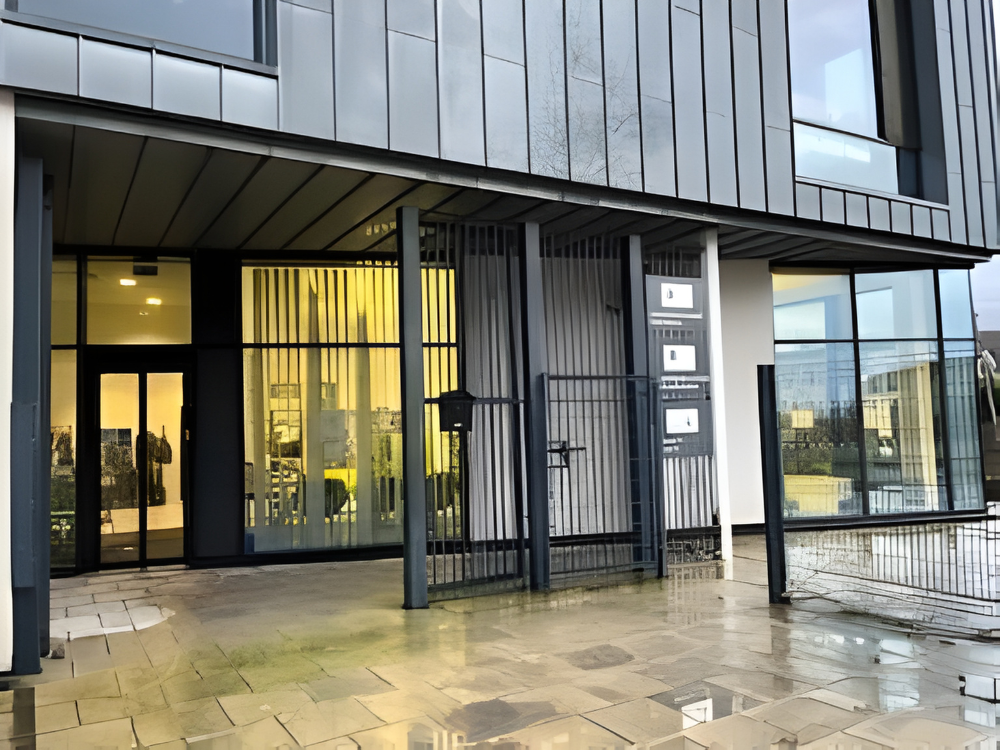 Modern building entrance with glass doors, tall metal bars, and large windows, featuring a bright interior visible from outside.