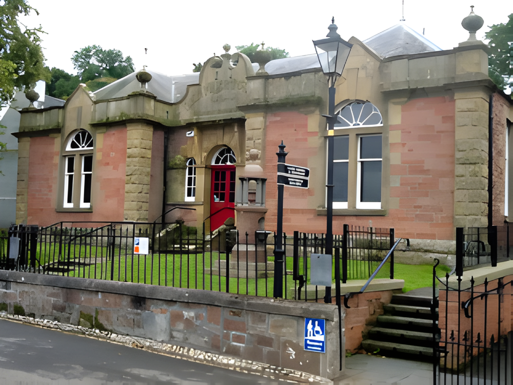 Historic stone library with arched windows, red door, iron fence, and street lamps in front.