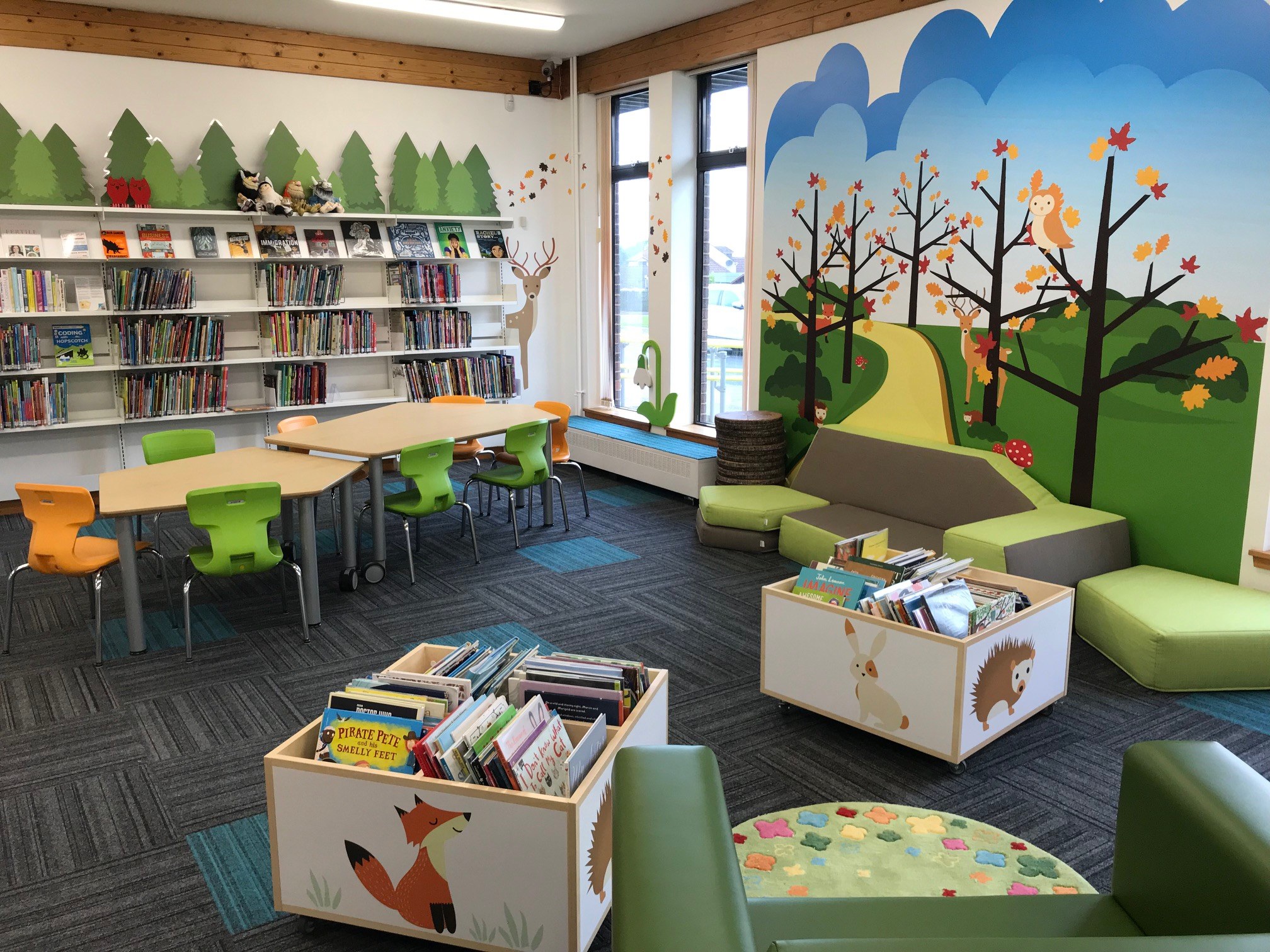 Children’s library area with colorful chairs, bookshelves, and a wall mural of trees and animals.